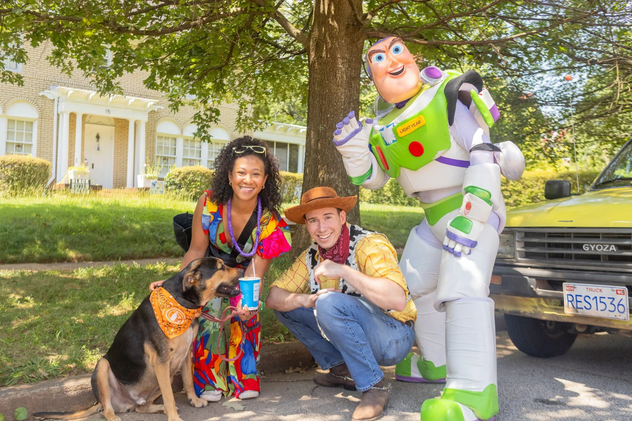 Two women and a man with a dog posing with a life-sized Buzz Lightyear statue on a sunny day in a residential neighborhood. The women are smiling, and the man is crouching down, holding a beverage. The dog is wearing an orange bandana, and the group 