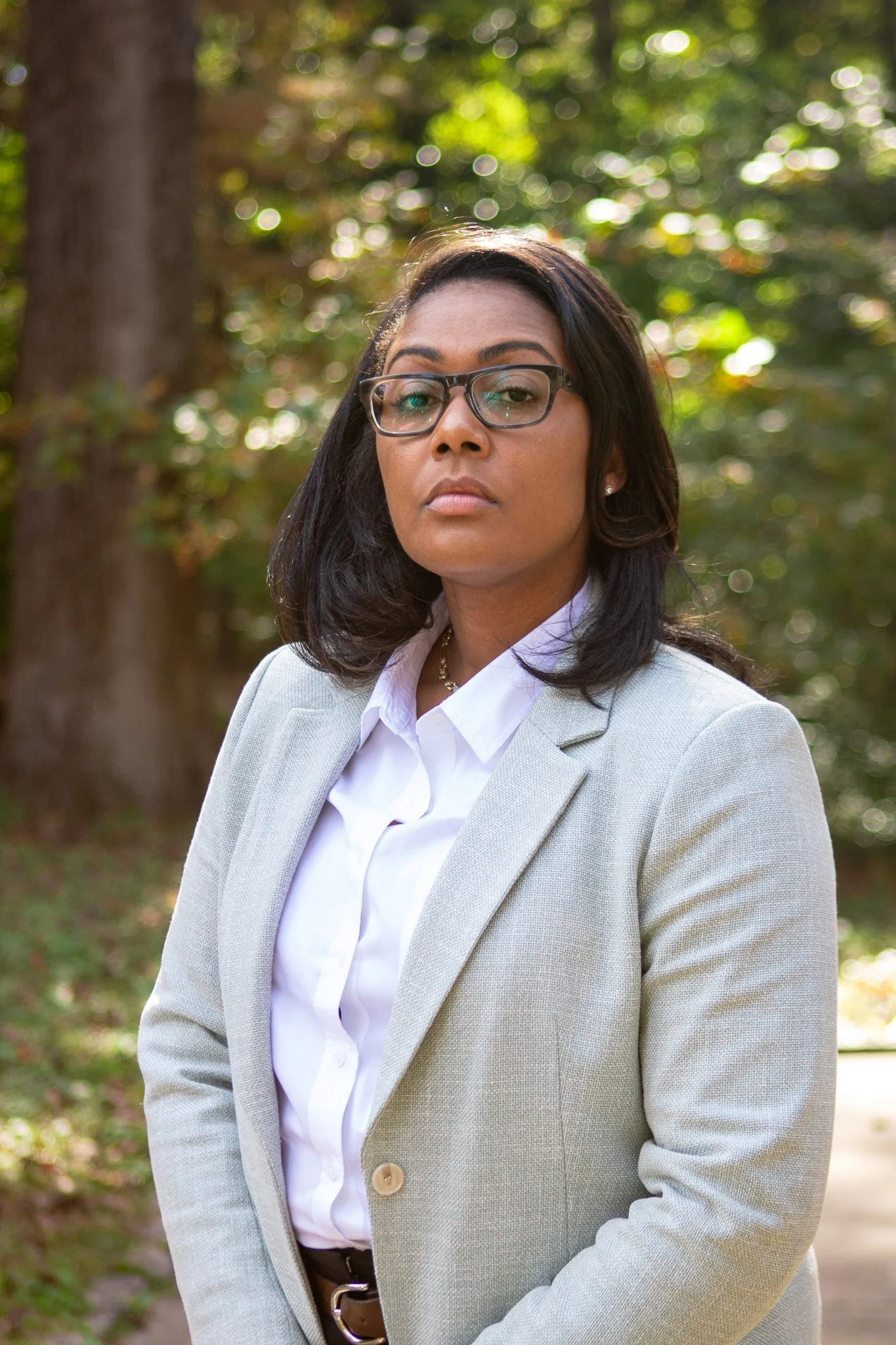 A woman wearing glasses and a light gray blazer standing outdoors with green foliage in the background.