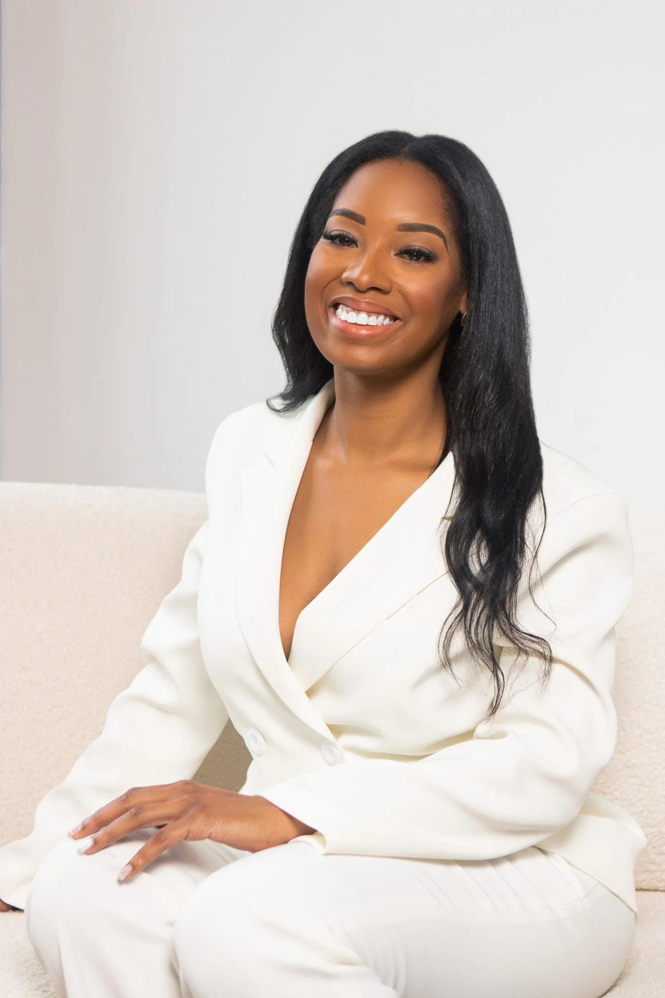 A woman with long black hair styled in loose waves, wearing a white suit, sitting on a beige couch against a plain light background, smiling and looking at the camera.