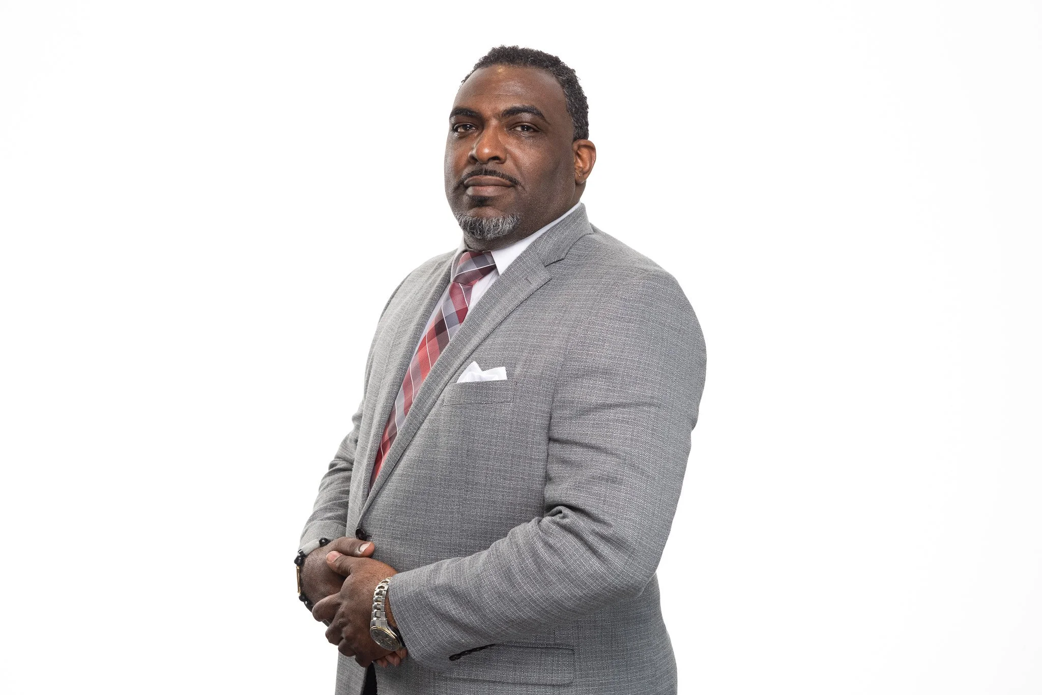 A man dressed in a gray suit with a white shirt and a striped tie, wearing a watch and a bracelet, standing against a plain white background.