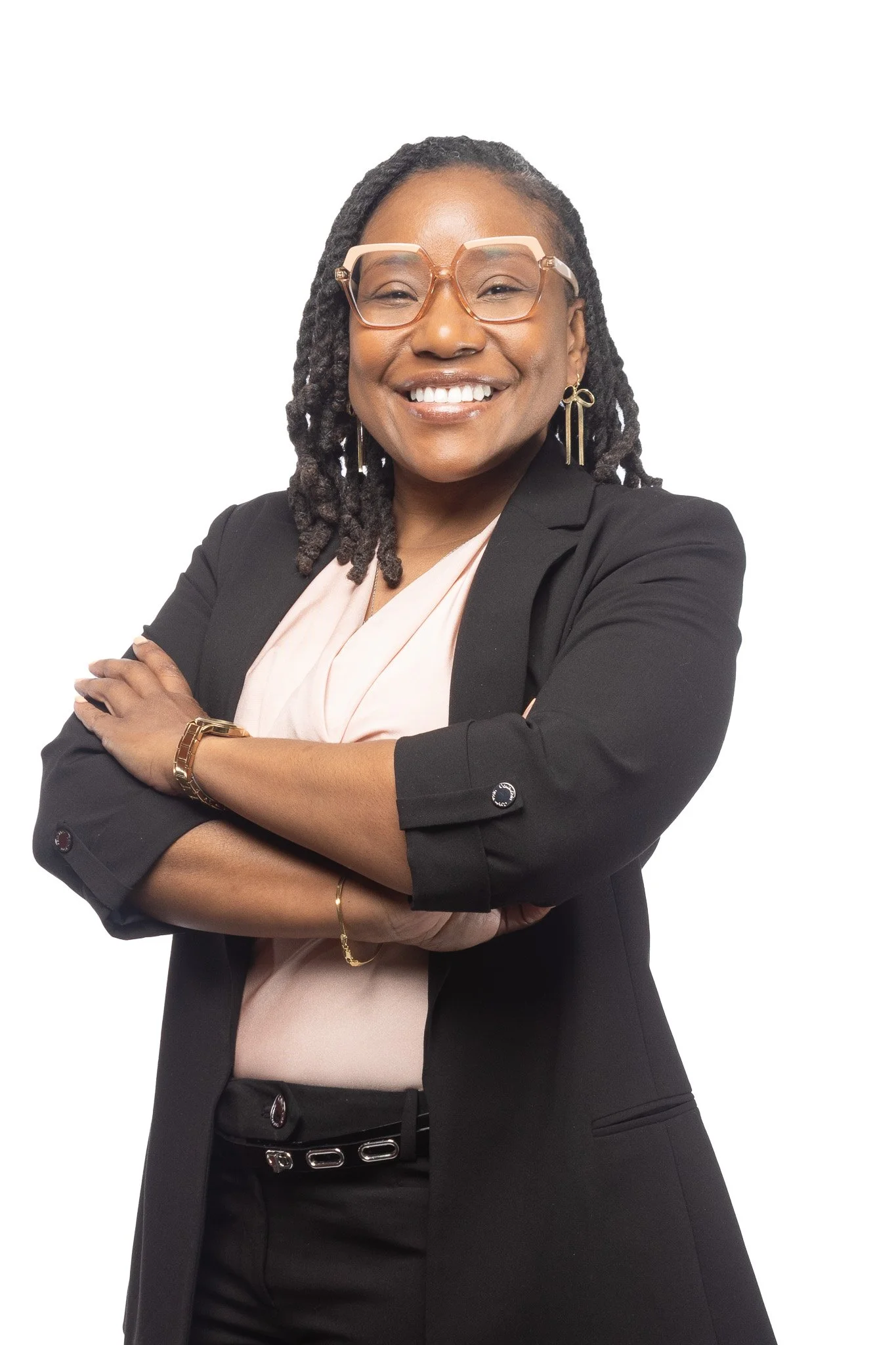 A smiling Black woman with glasses, wearing a black blazer, pale pink blouse, and gold jewelry, standing with crossed arms against a white background.