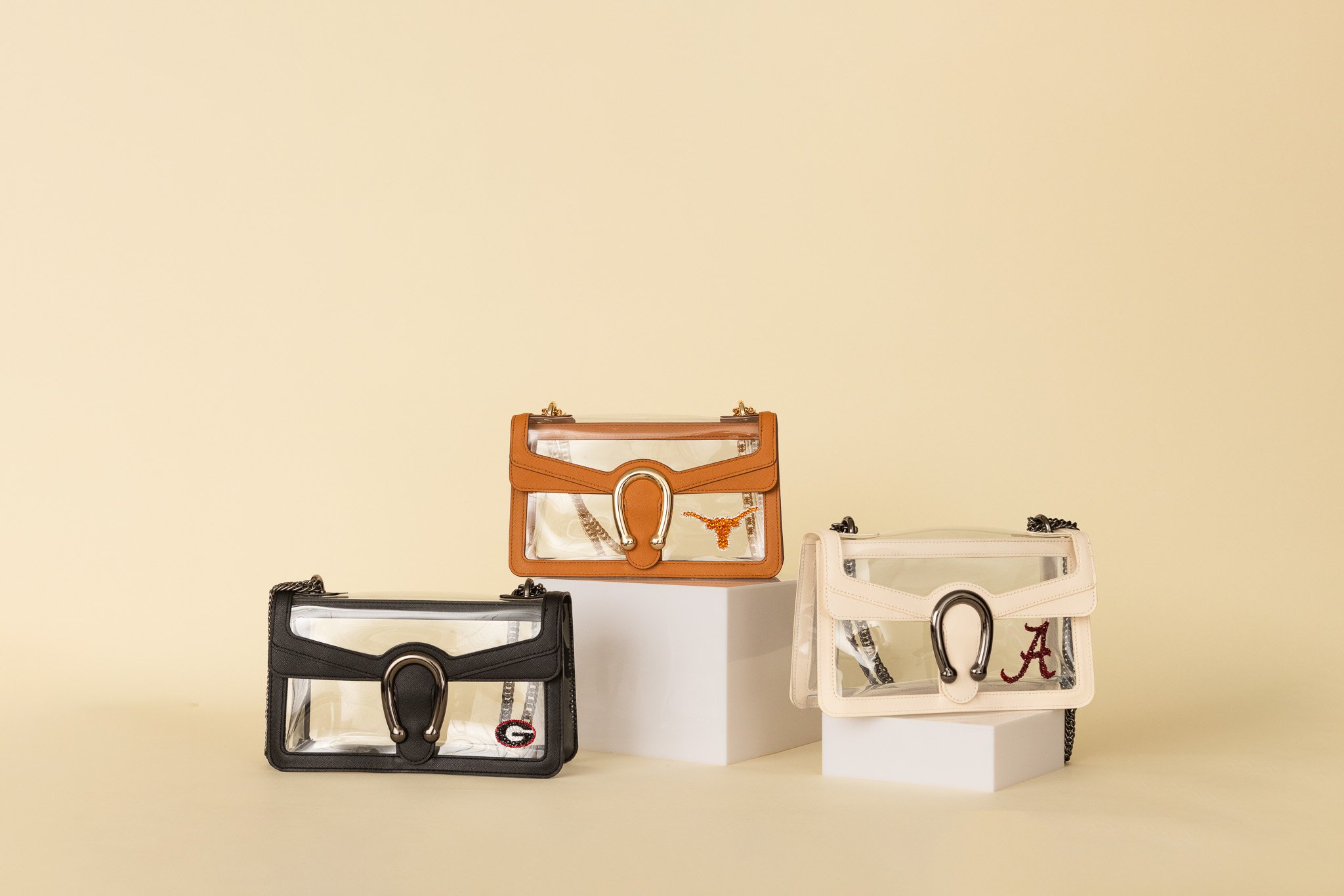 Four designer handbags in black, tan, white, and clear plastic with chain straps, displayed on white platforms against a beige background.