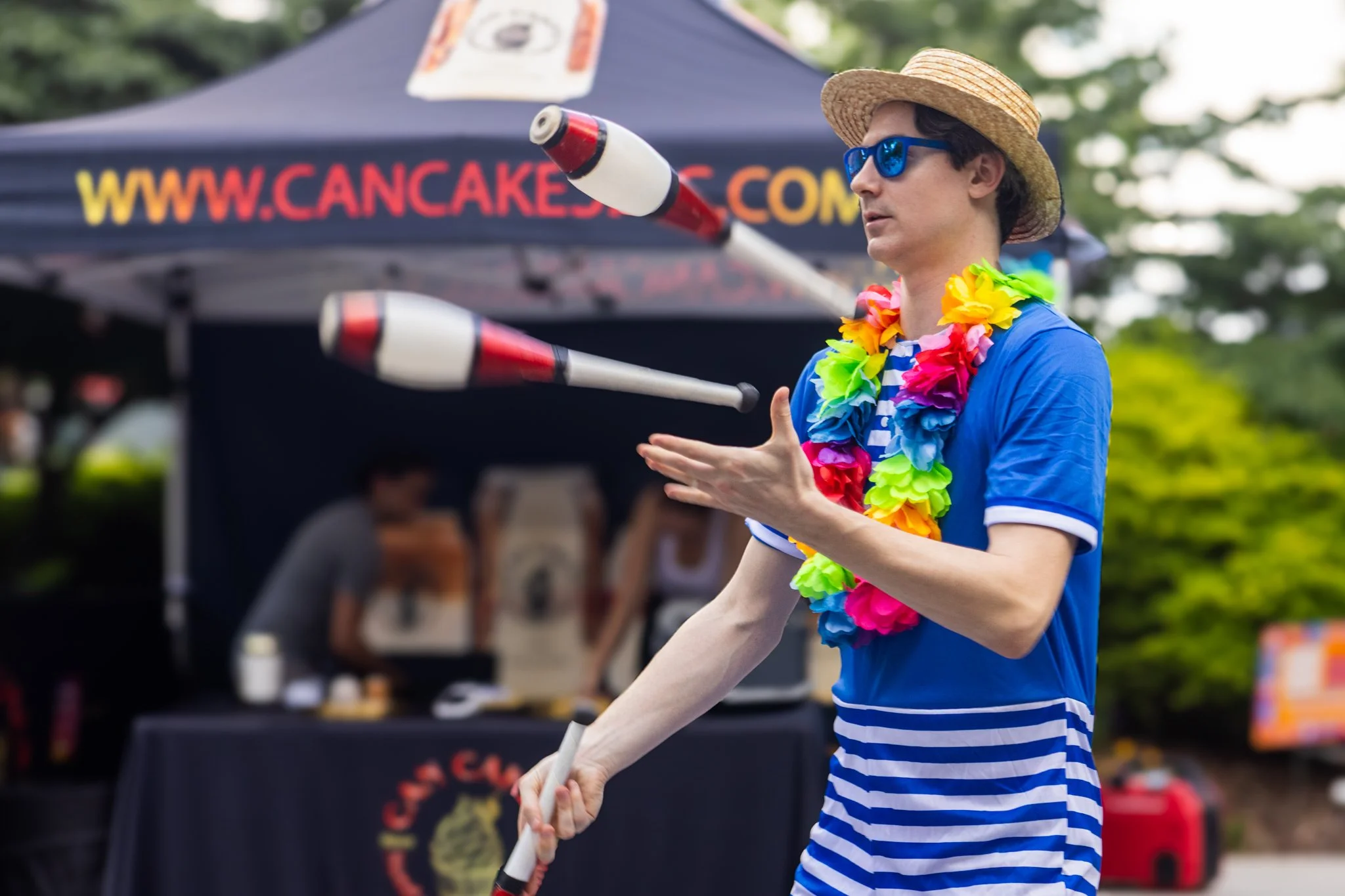 Person in a blue and white striped shirt and a straw hat playing juggling balls, wearing a colorful lei, at an outdoor event with a canopy tent in the background.