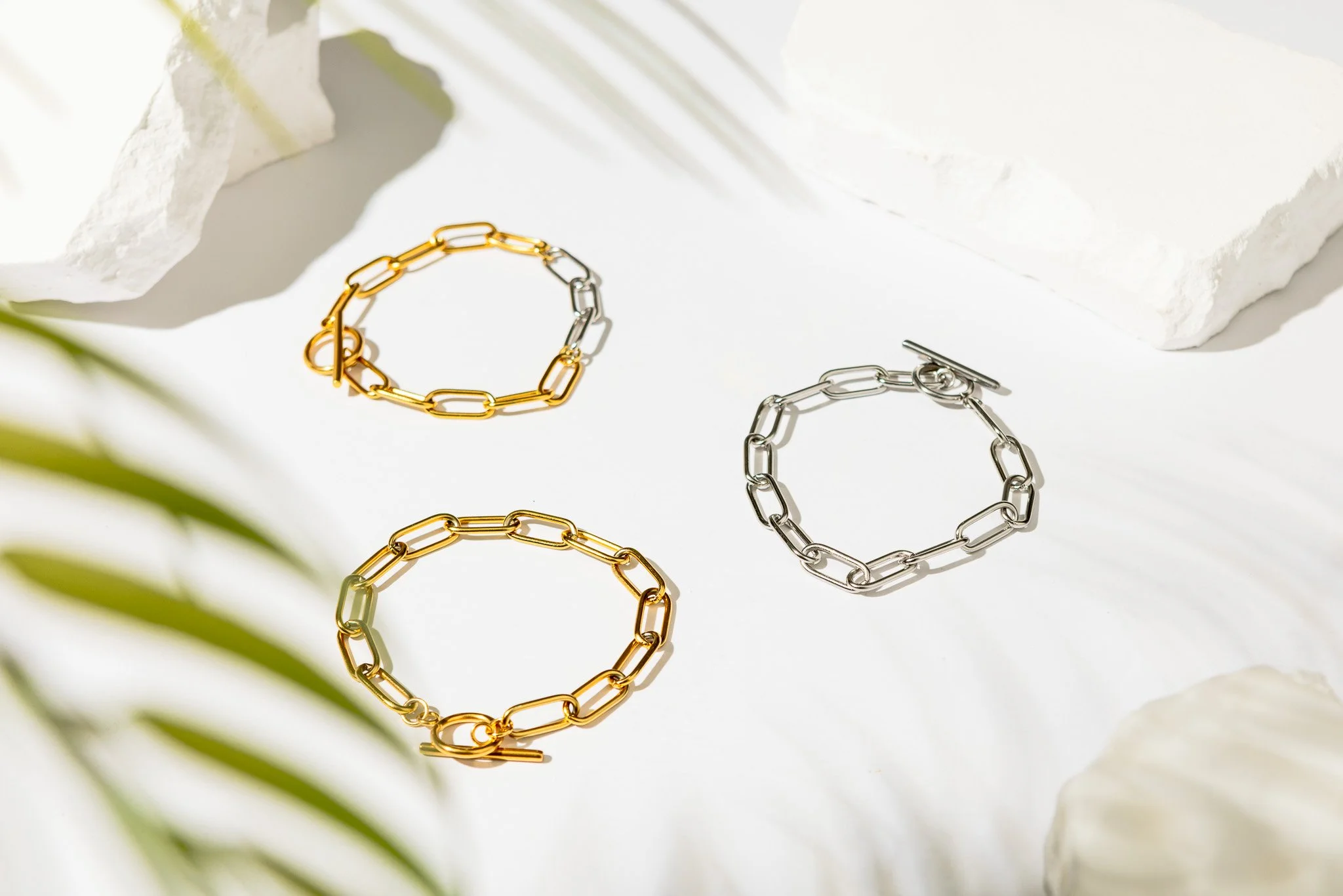 Gold and silver chain link bracelets on a white surface with rocks and greenery.