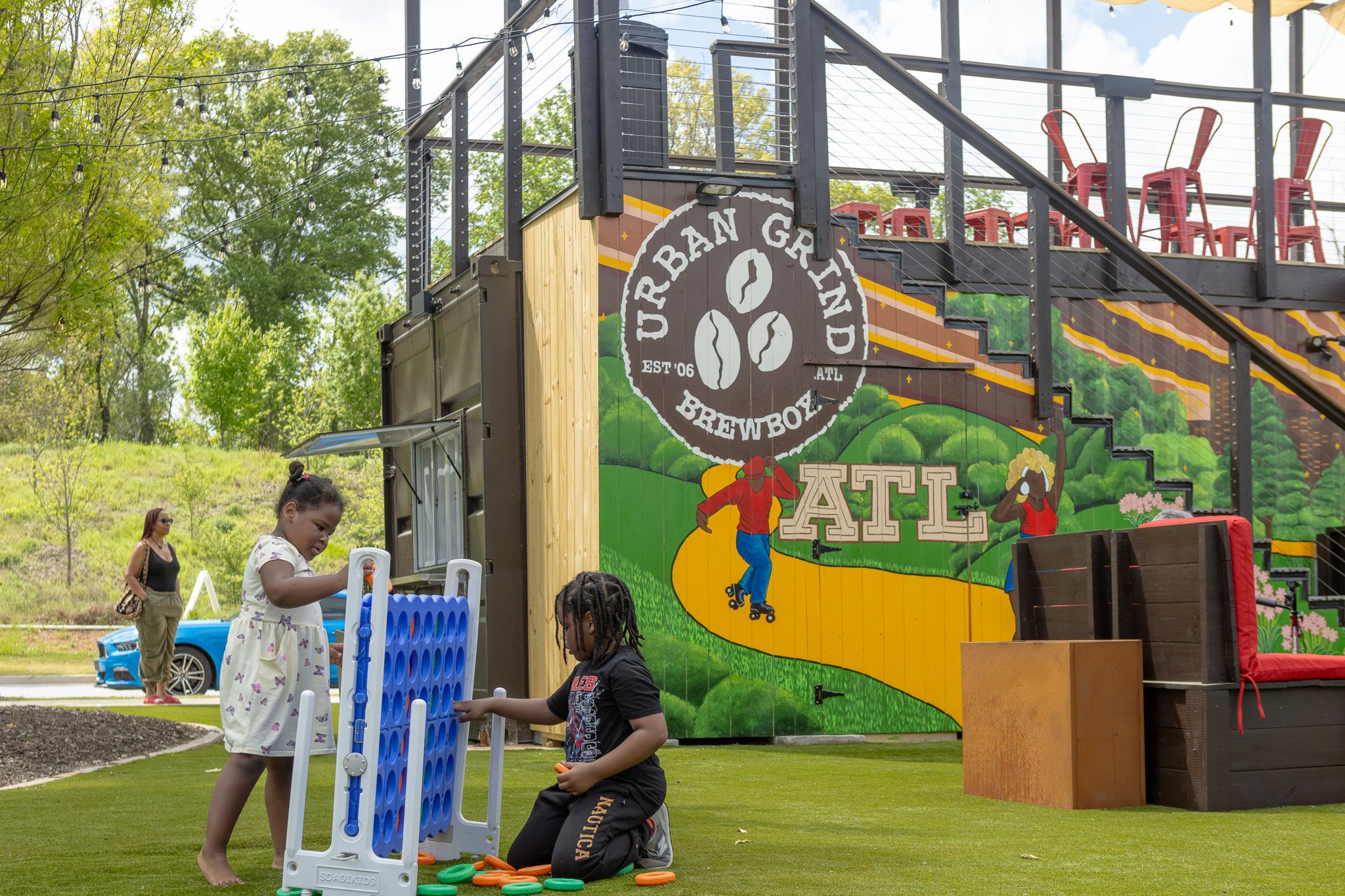 Children playing a game of Connect Four outdoors on a grassy area with trees and a building with colorful artwork in the background that promotes a brewery. Some adults are visible in the background, and there are red chairs on a deck above.