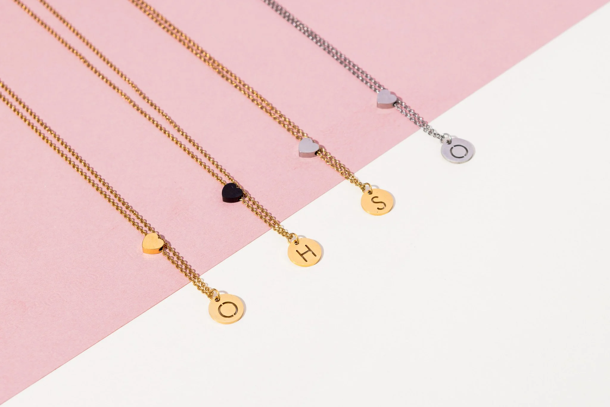 Four delicate necklaces with heart and initial charms, in gold and silver, arranged on a pink and white surface.