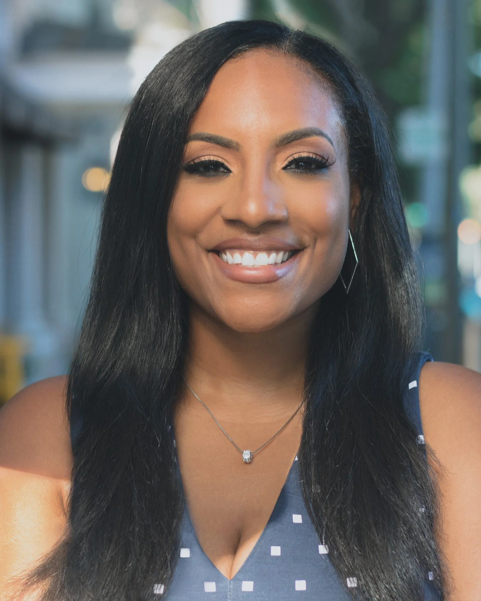 Close-up of a smiling woman with long black hair, wearing a navy blue dress with white polka dots, earrings, and a pendant necklace.