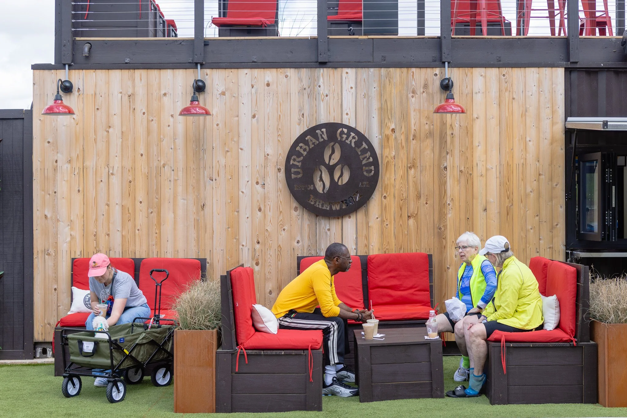 People sitting on red cushioned outdoor seating and socializing in front of a wooden wall with a logo that says 'Urban Green'.