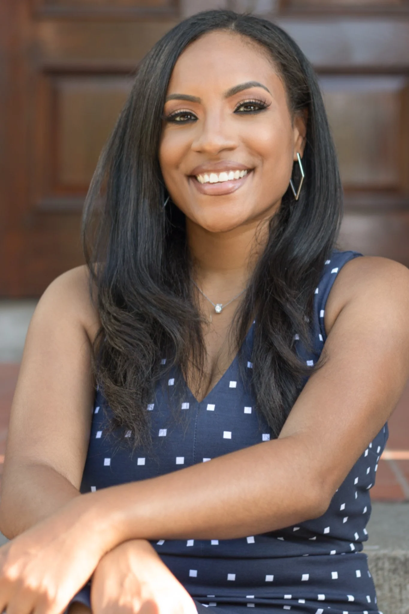 A woman with long dark hair, wearing a navy sleeveless dress with white square pattern, smiling, with earrings and a necklace, sitting outdoors.