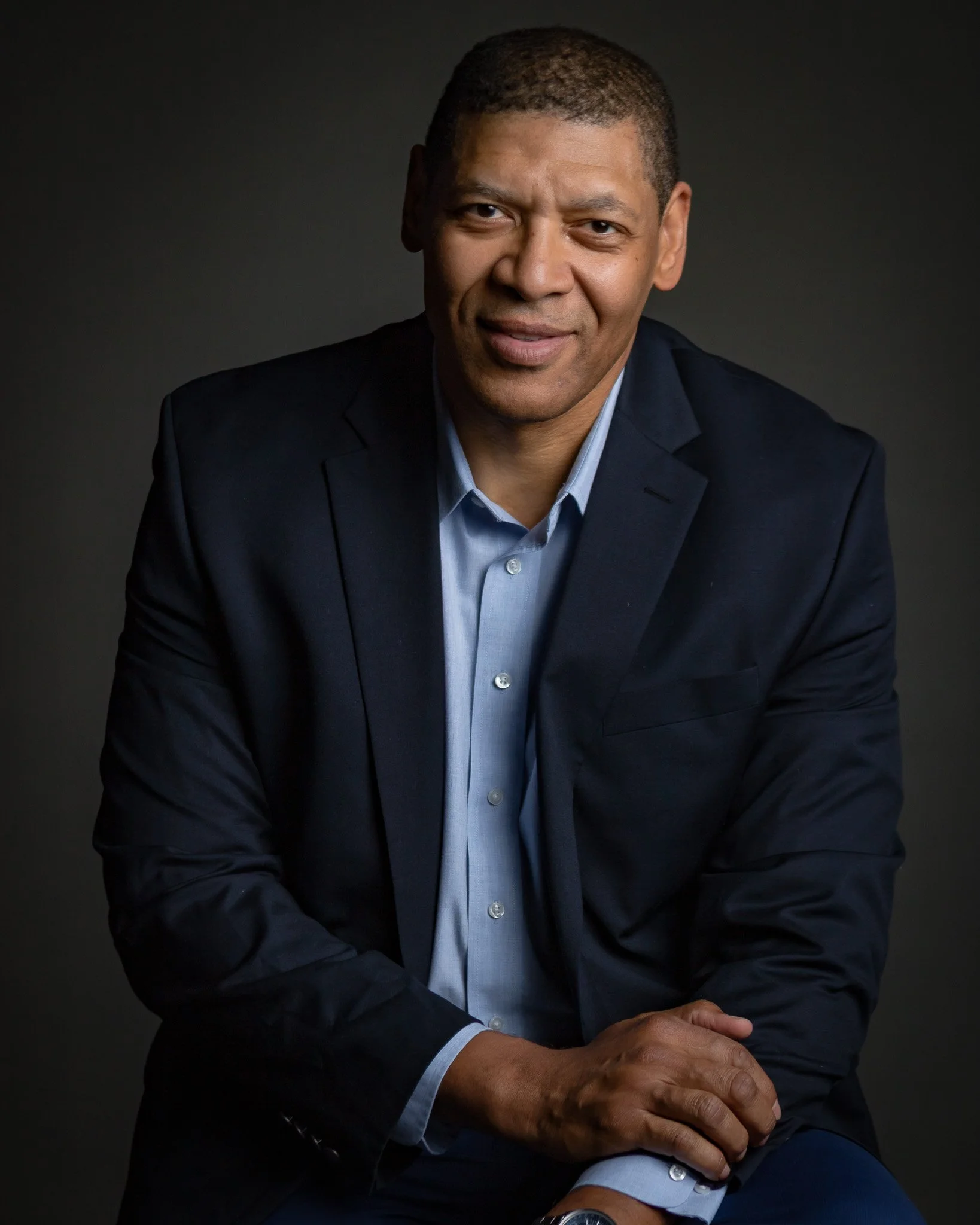 Portrait of a man with short curly hair, wearing a navy blazer and light blue shirt, sitting with his hands clasped, against a dark background.
