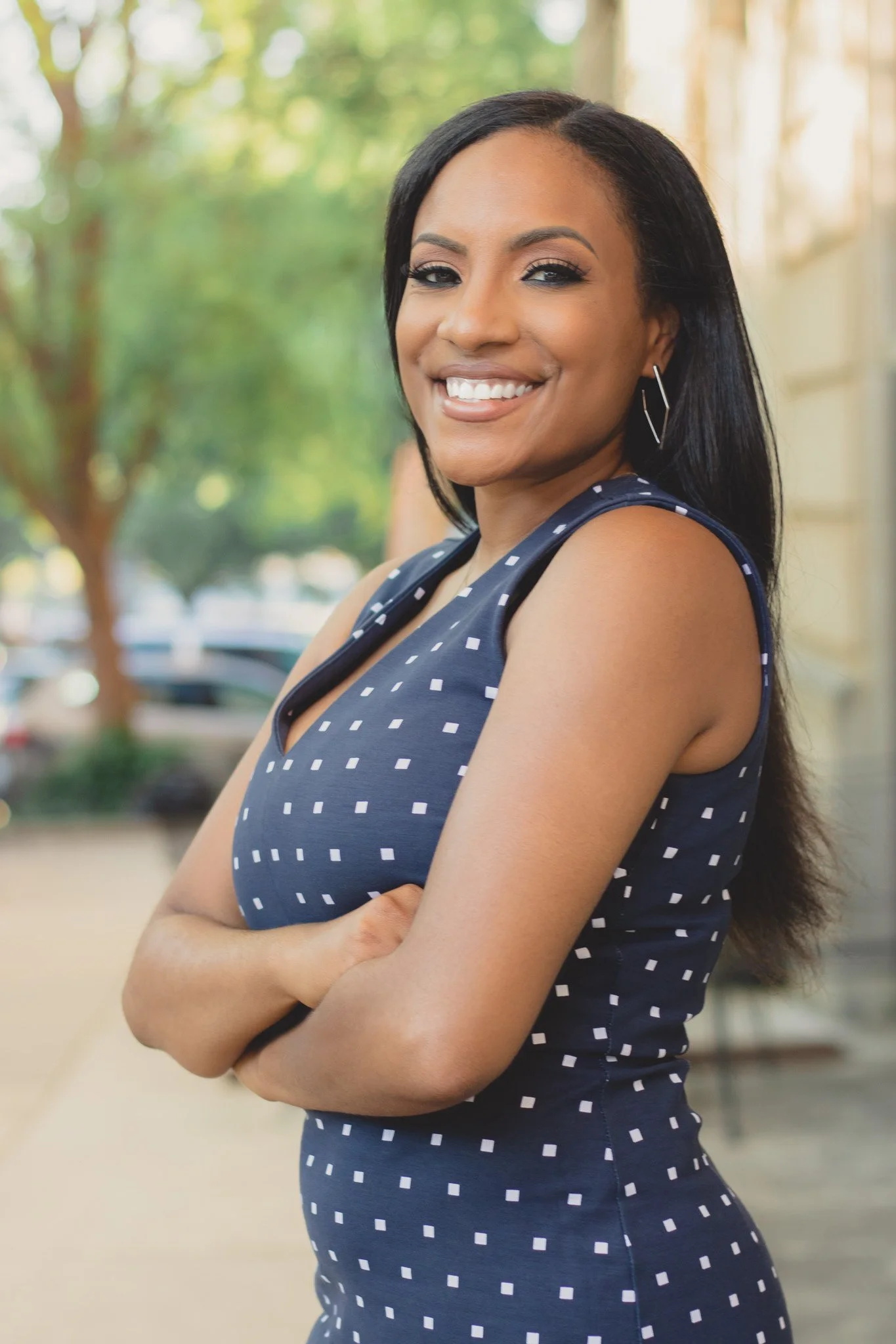 A woman with long black hair and hoop earrings smiling with arms crossed, wearing a dark blue sleeveless dress with white squares, outdoors with trees and sunlight in the background.