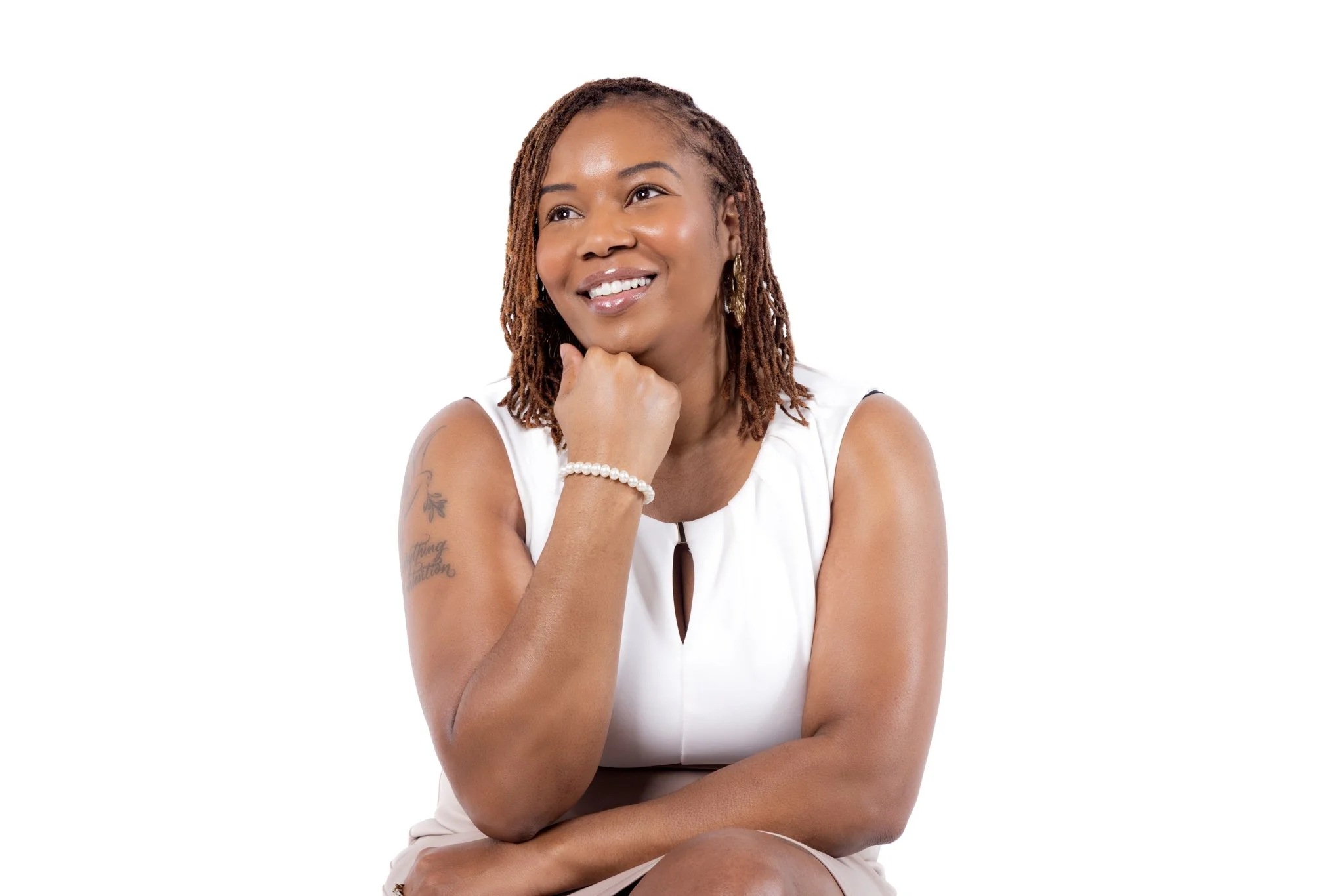 A woman with medium brown skin and red dreadlocks is smiling and looking up. She is resting her chin on her hand and wearing a white sleeveless top, a pearl bracelet, and earrings. She has a tattoo on her left arm.
