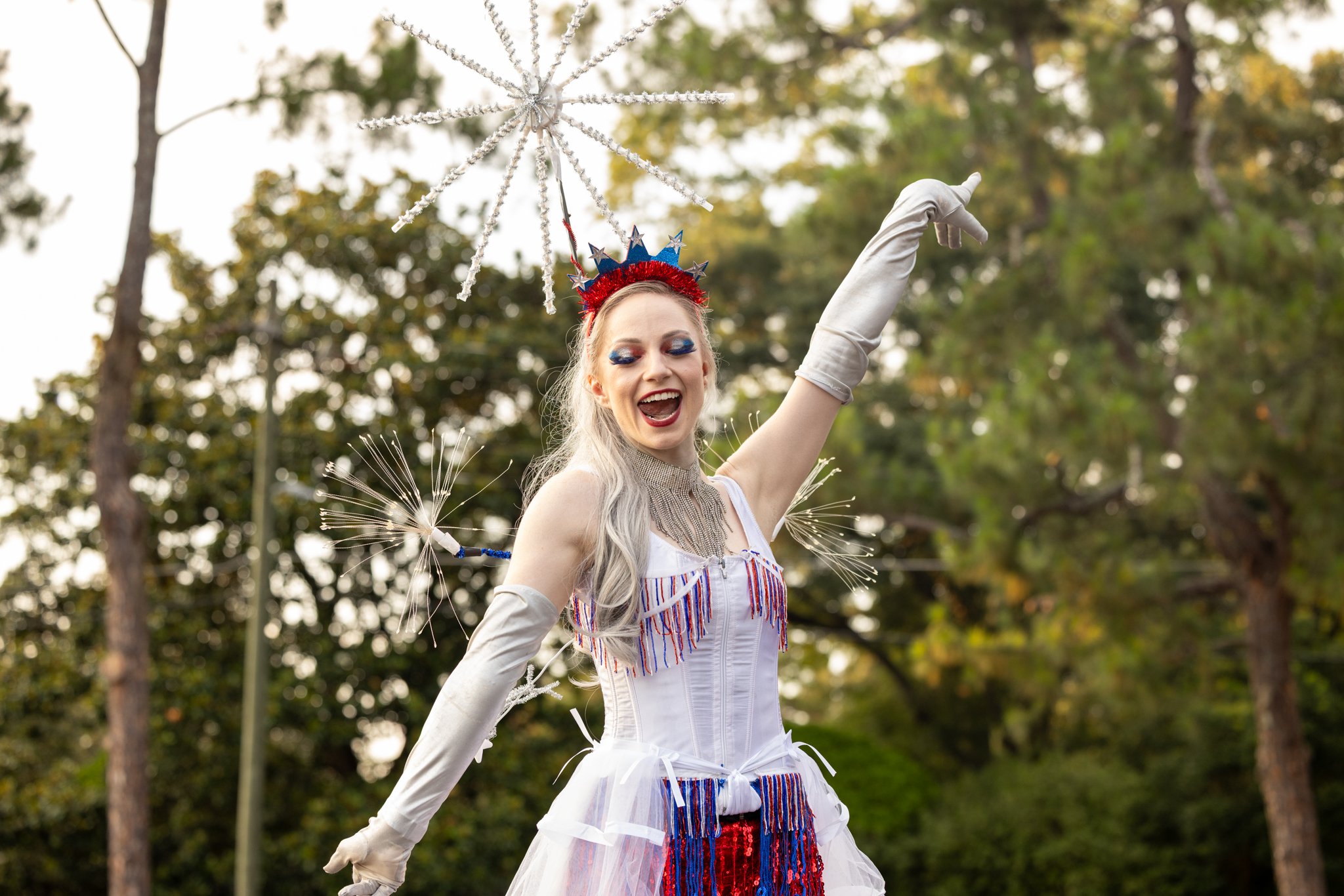 A woman smiling and dressed in American flag-themed costume with patriotic accessories, waving outdoors in a wooded area during daylight.