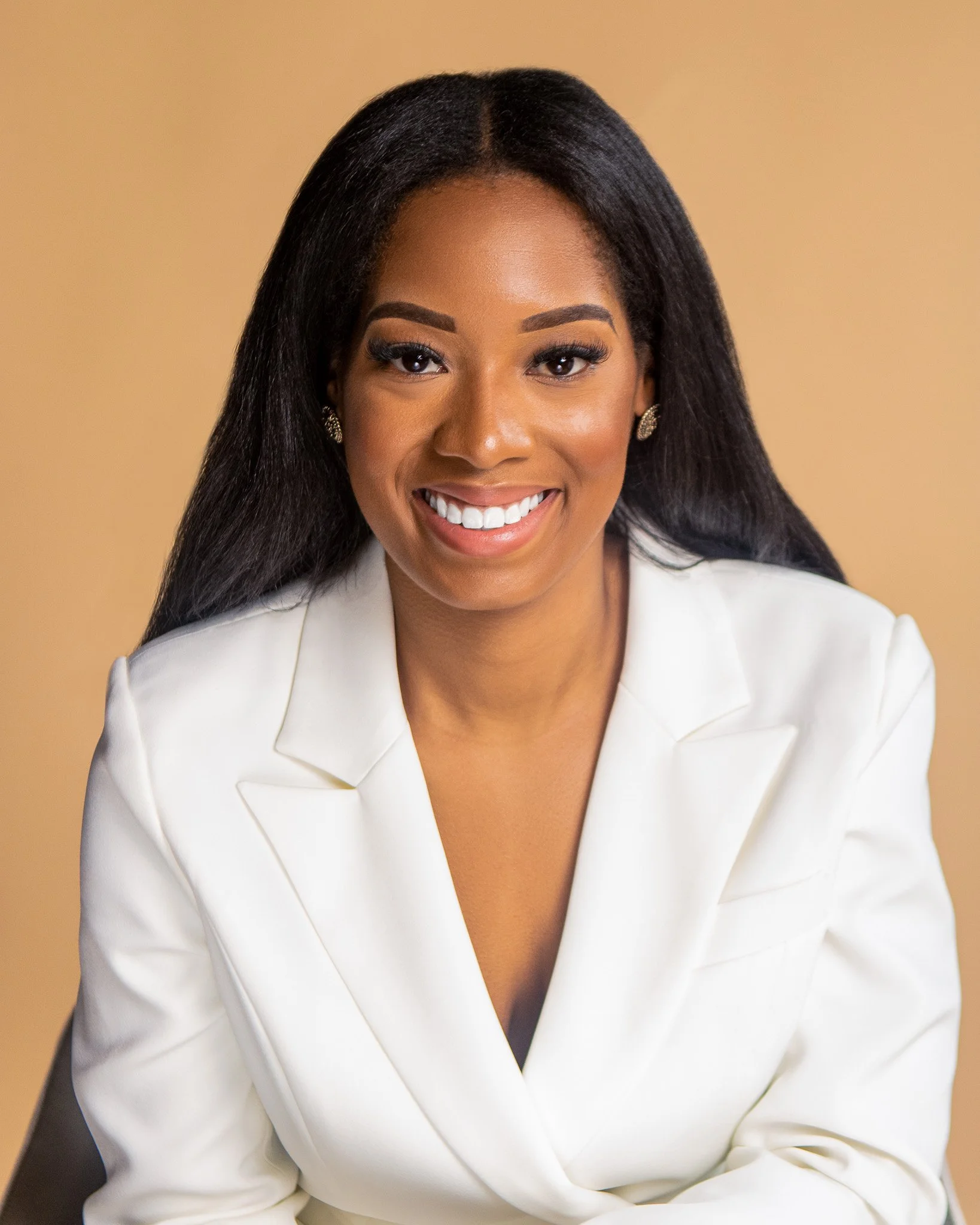 Close-up portrait of an African American woman smiling, wearing a white blazer, with black hair styled straight, set against a beige background.