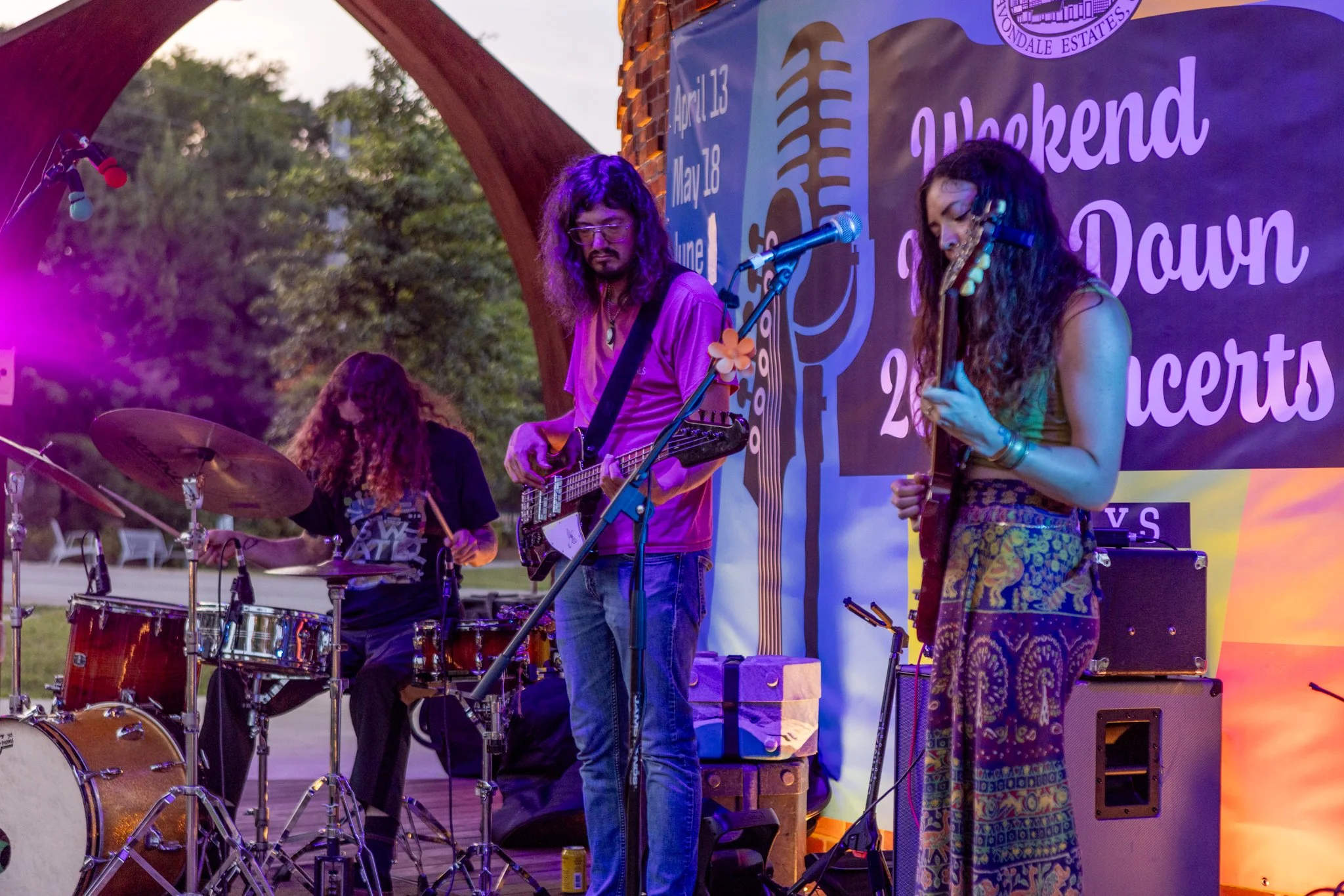 Musicians performing on an outdoor stage at dusk. A drummer, a guitarist, and a female singer are visible, with a colorful backdrop featuring event details.