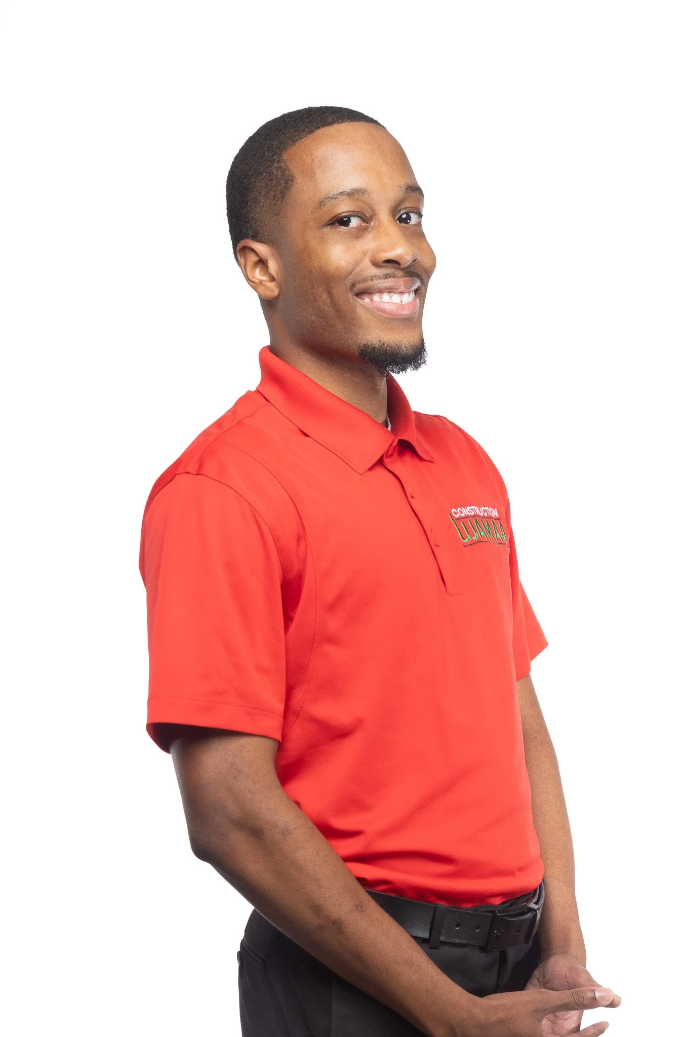 A smiling young man wearing a red polo shirt and black pants, with a belt, and standing against a white background.