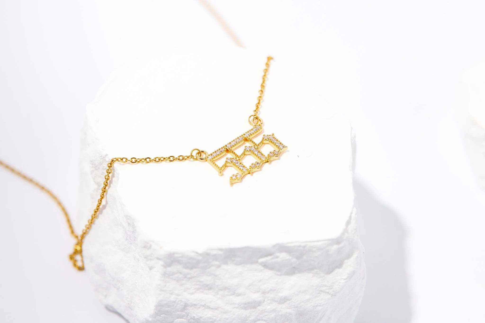 Gold necklace with a pendant that features the word 'beach' in stylized letters, decorated with small gemstones, placed on a white textured surface.