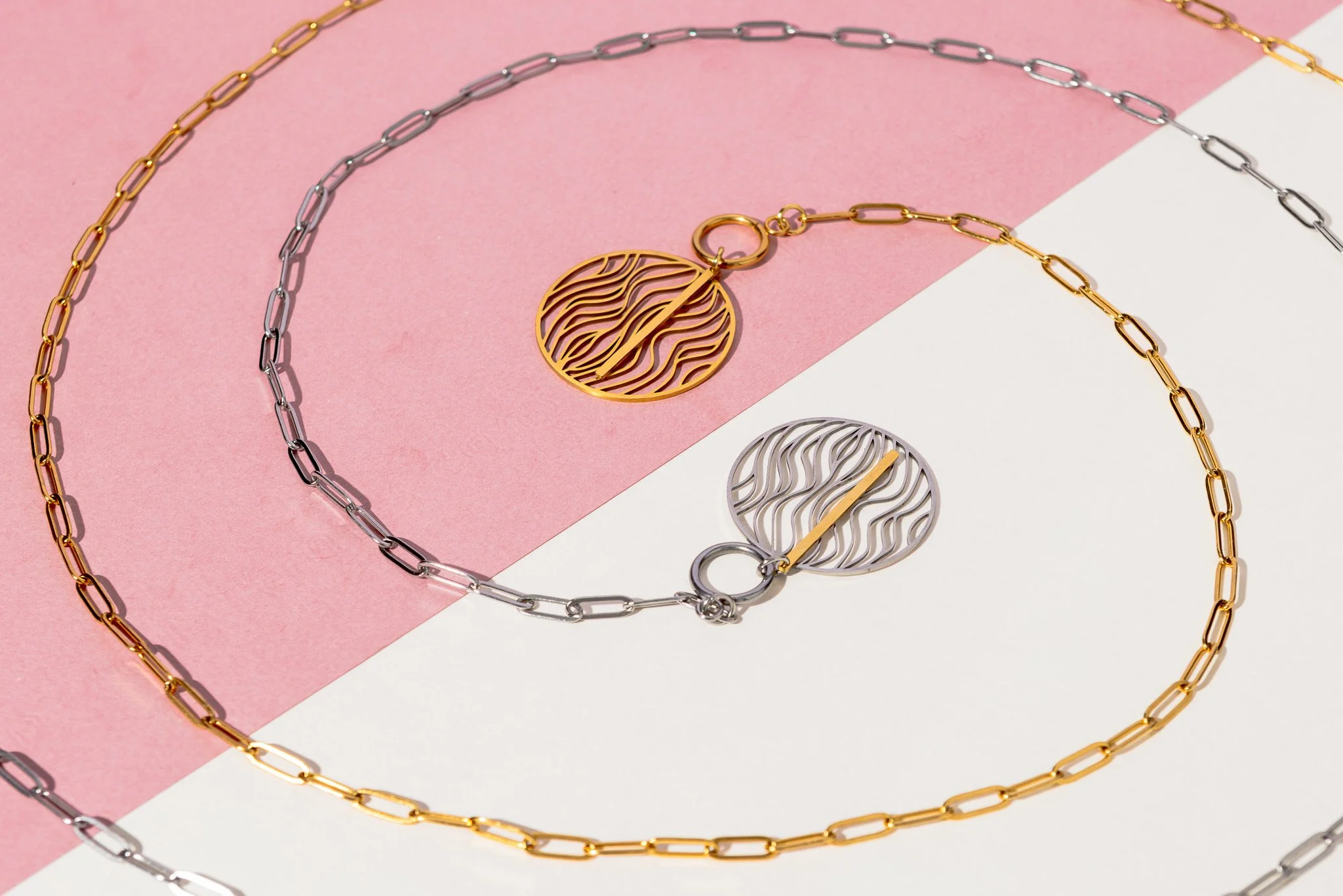 Gold and silver chain necklaces with round, filigree pendant charms on a pink and white background.
