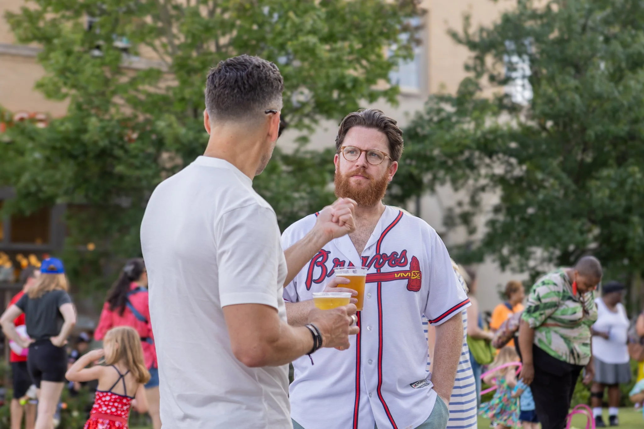 Two men with glasses are standing and talking at an outdoor event, holding plastic cups of beer. Visible are a crowd of people, a child in a red polka dot dress, and trees in the background.
