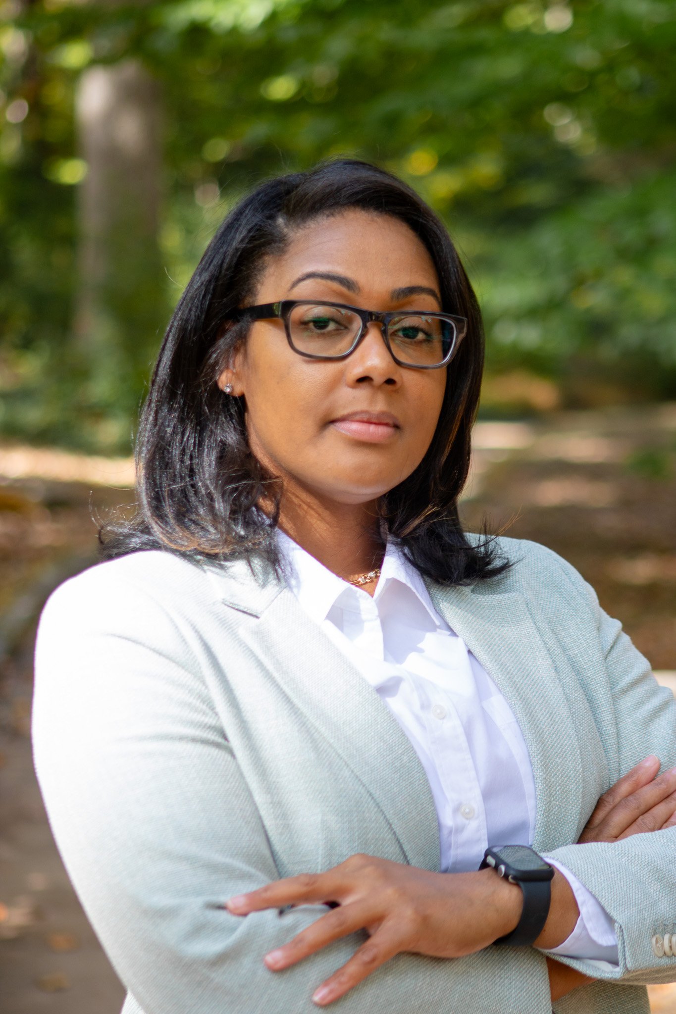 A woman with black hair and glasses wearing a light gray blazer over a white shirt, standing outdoors with blurred green trees in the background.