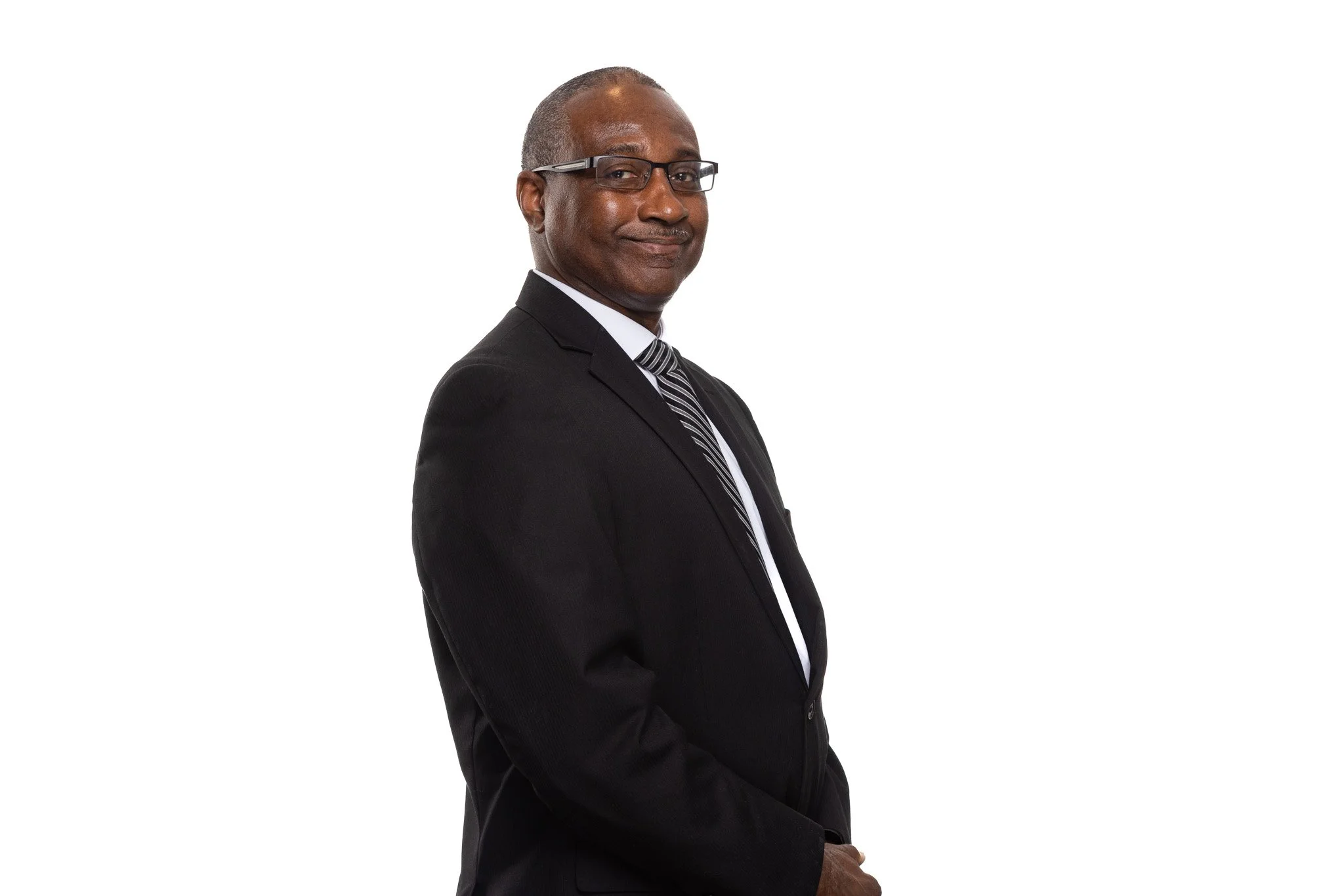 An older man with gray hair and glasses, dressed in a black suit, white shirt, and striped tie, stands against a white background with a confident expression.