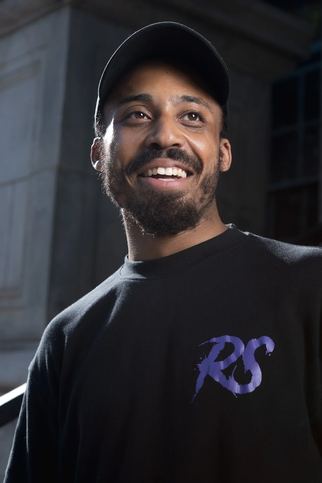 Close-up of a smiling man with a beard and mustache wearing a black cap and black T-shirt with purple graphic, outdoors in natural light.