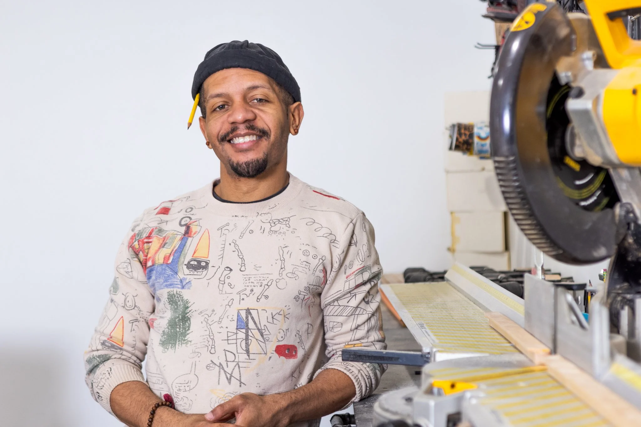 A man with a beard, wearing a black beanie and a light-colored sweater with colorful abstract designs, smiling while standing in a workshop near a power tool and workbench.