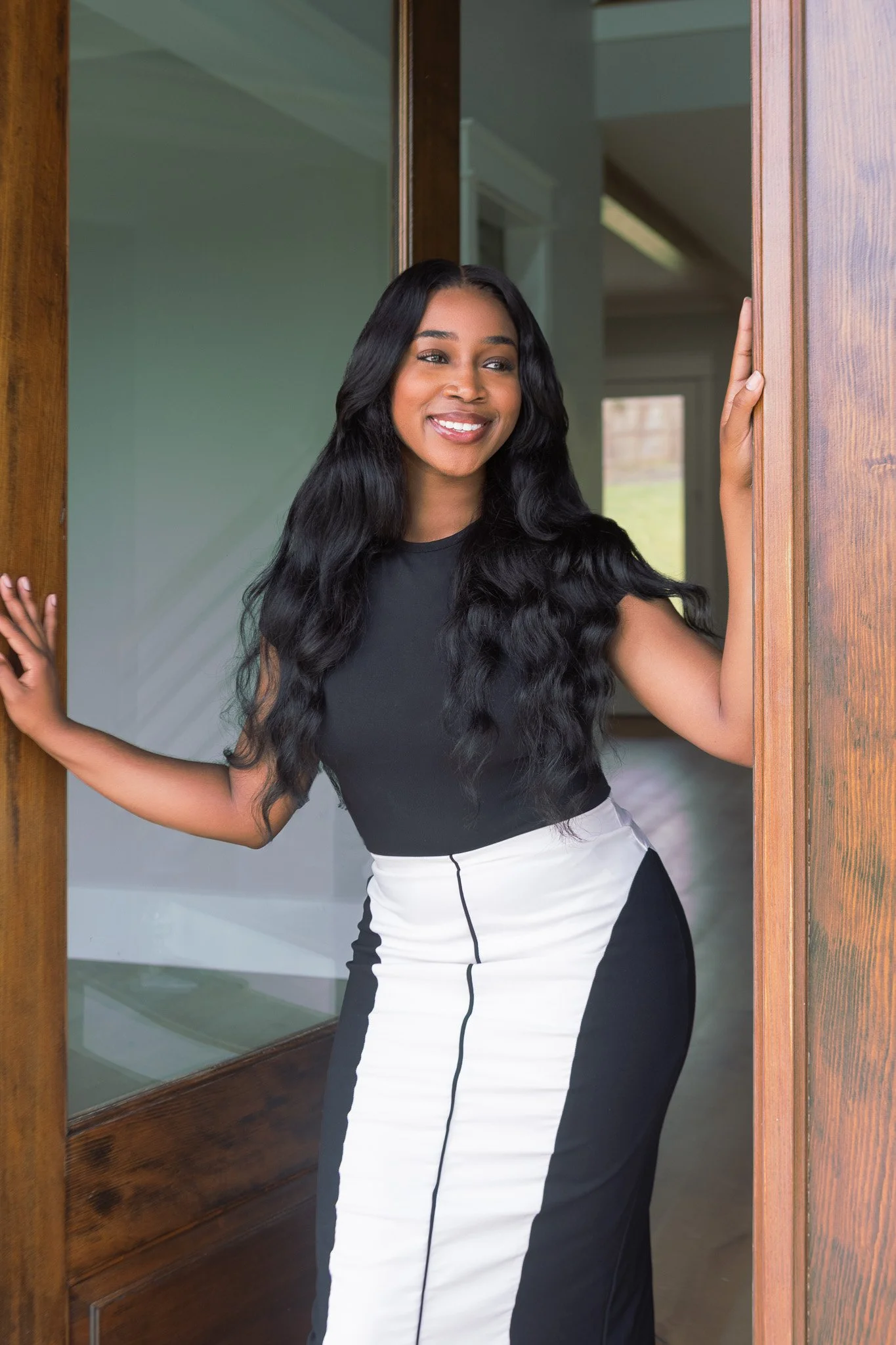 A woman with long black wavy hair smiling and standing by a wooden door, wearing a black and white color-block dress.