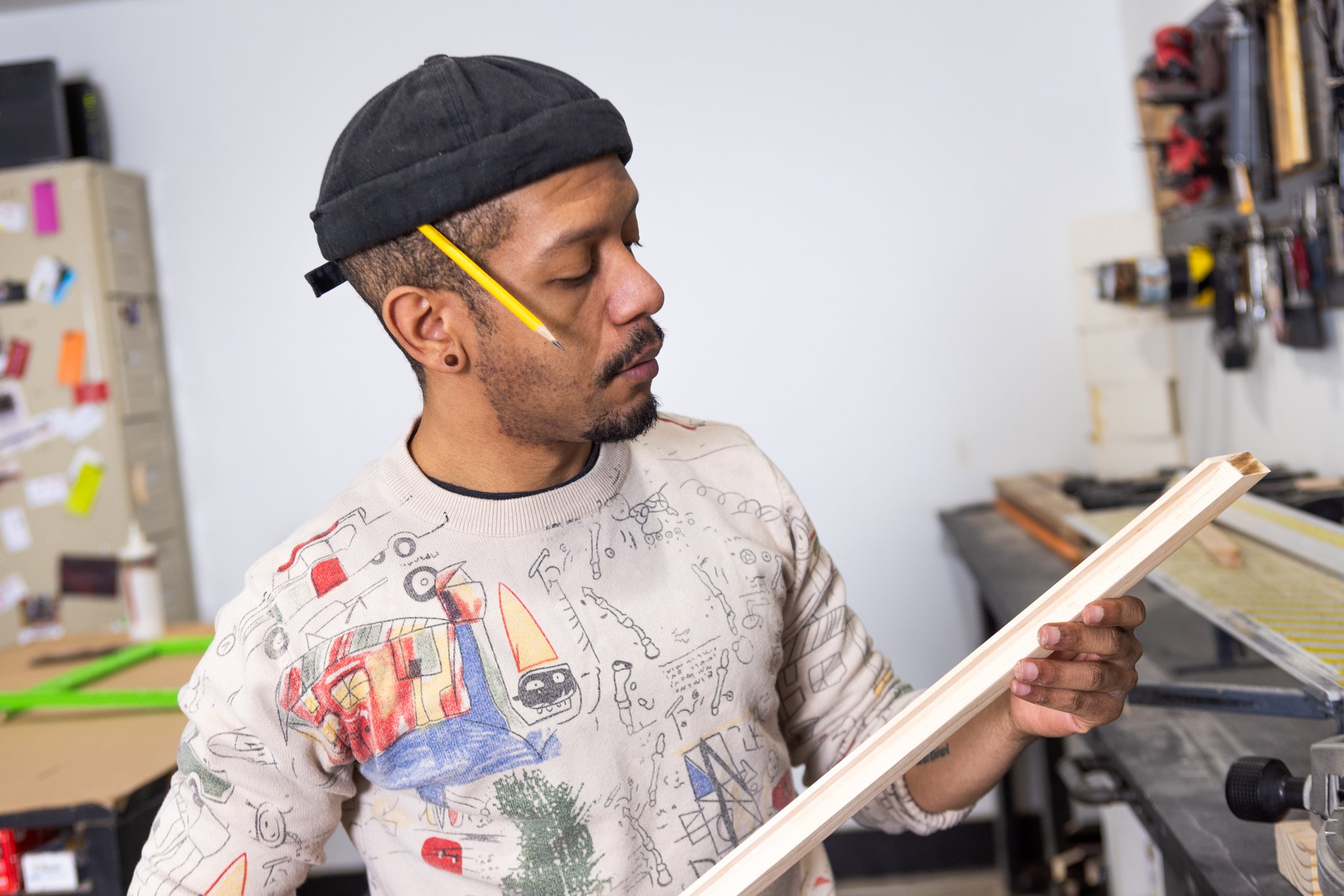 A man with a black beanie looking at a piece of wood in a workshop.