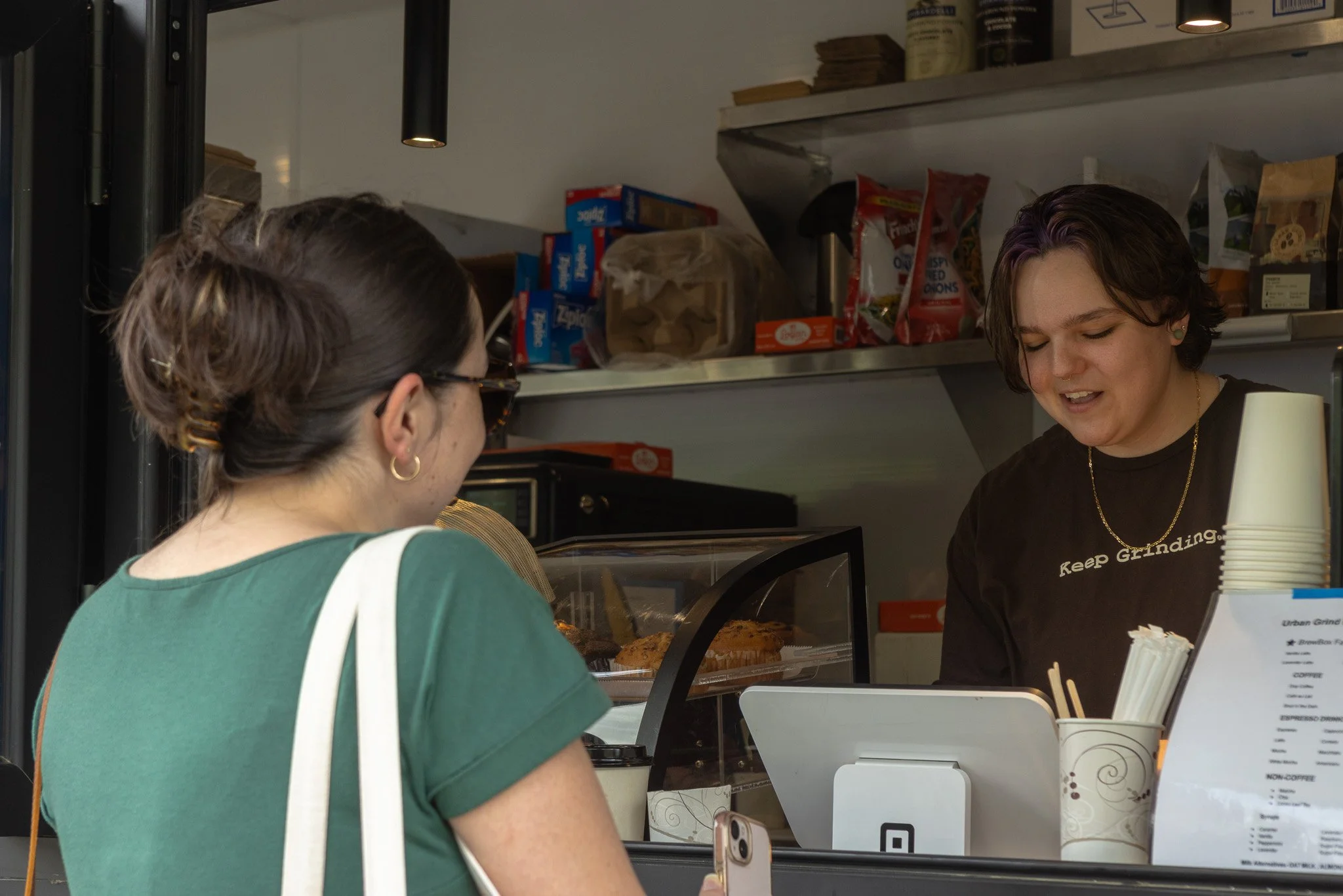 A woman with short purple and brown hair wearing a black shirt with white text, smiling and talking to another woman with glasses and a bun, at a counter with food items in a cafe or bakery.