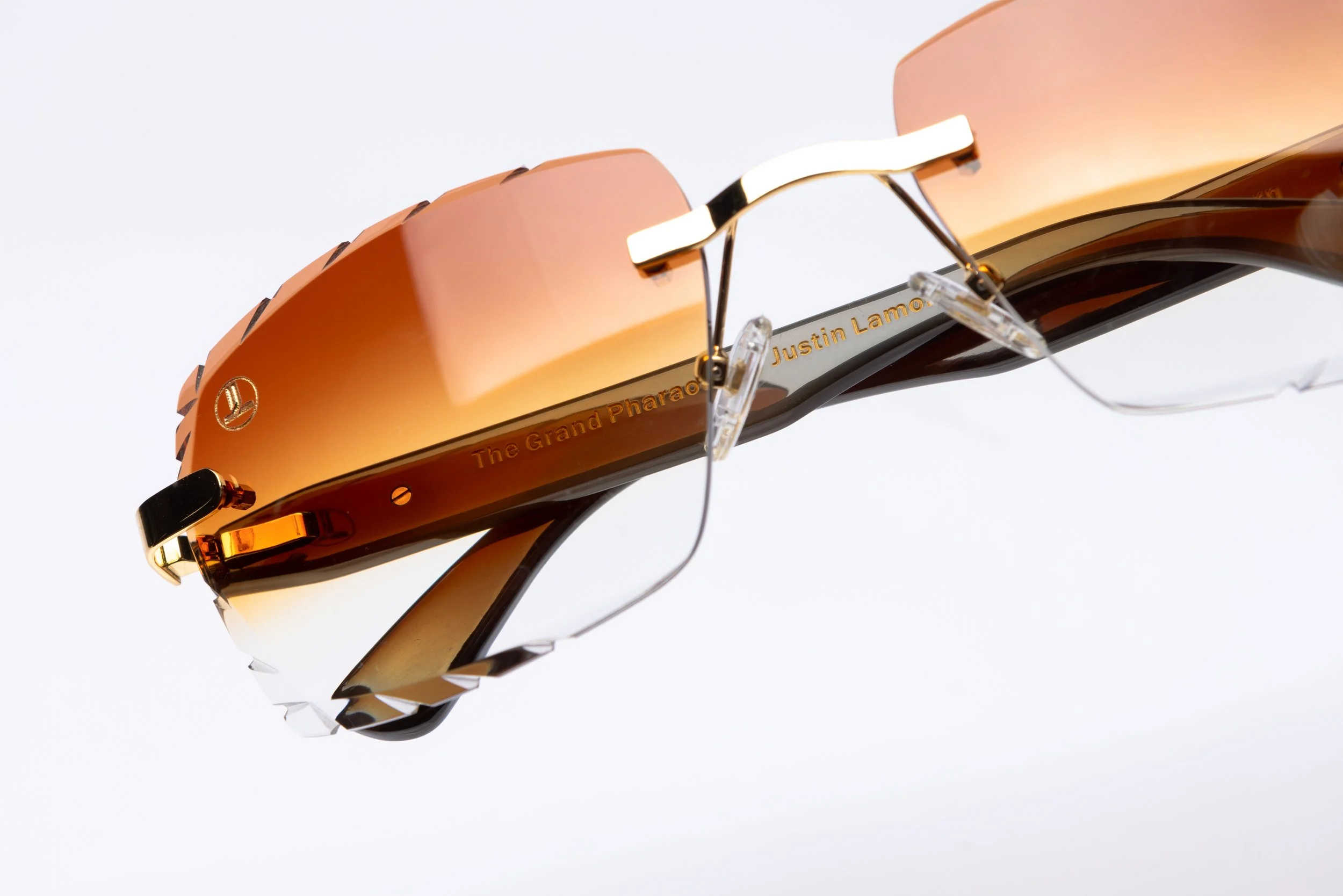 A pair of sunglasses with gold and brown lenses, featuring engraved text 'The Grand Pharaoh Justin Lamor' on the inside of the frame.