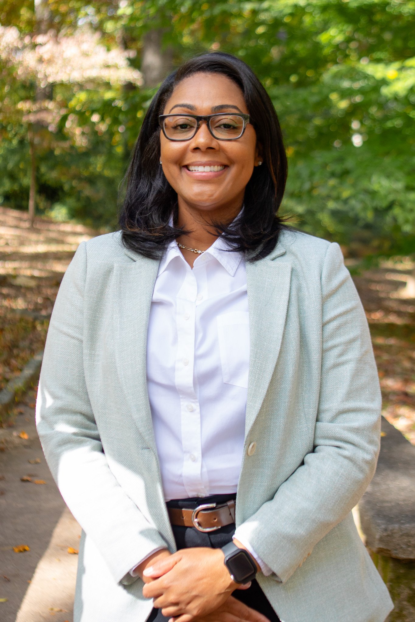 A woman with dark hair, glasses, and a light gray blazer standing outdoors on a sidewalk, smiling with trees and foliage in the background.