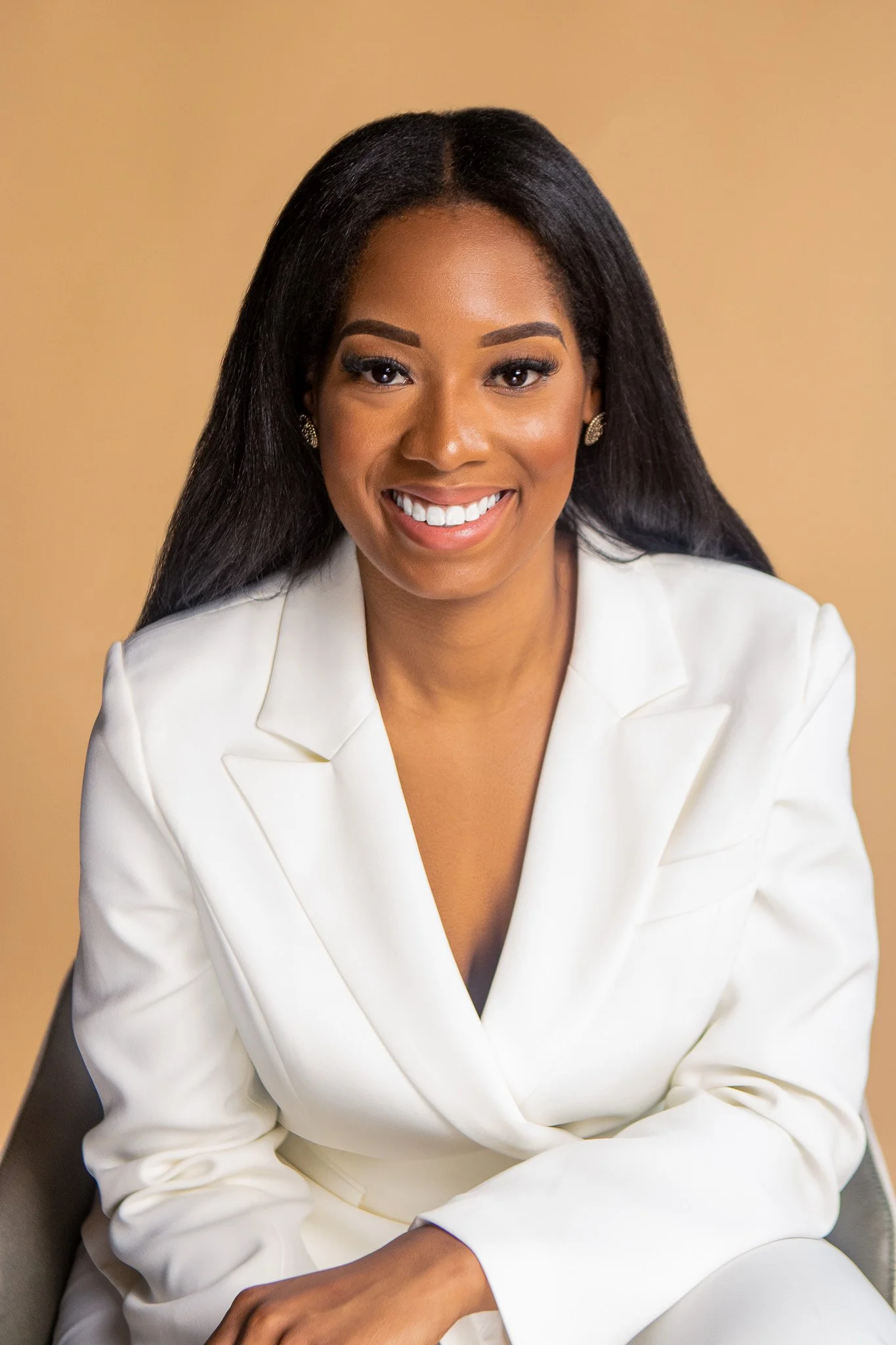 Portrait of a smiling African American woman with long black hair, wearing a white blazer and gold earrings, sitting against a beige background.