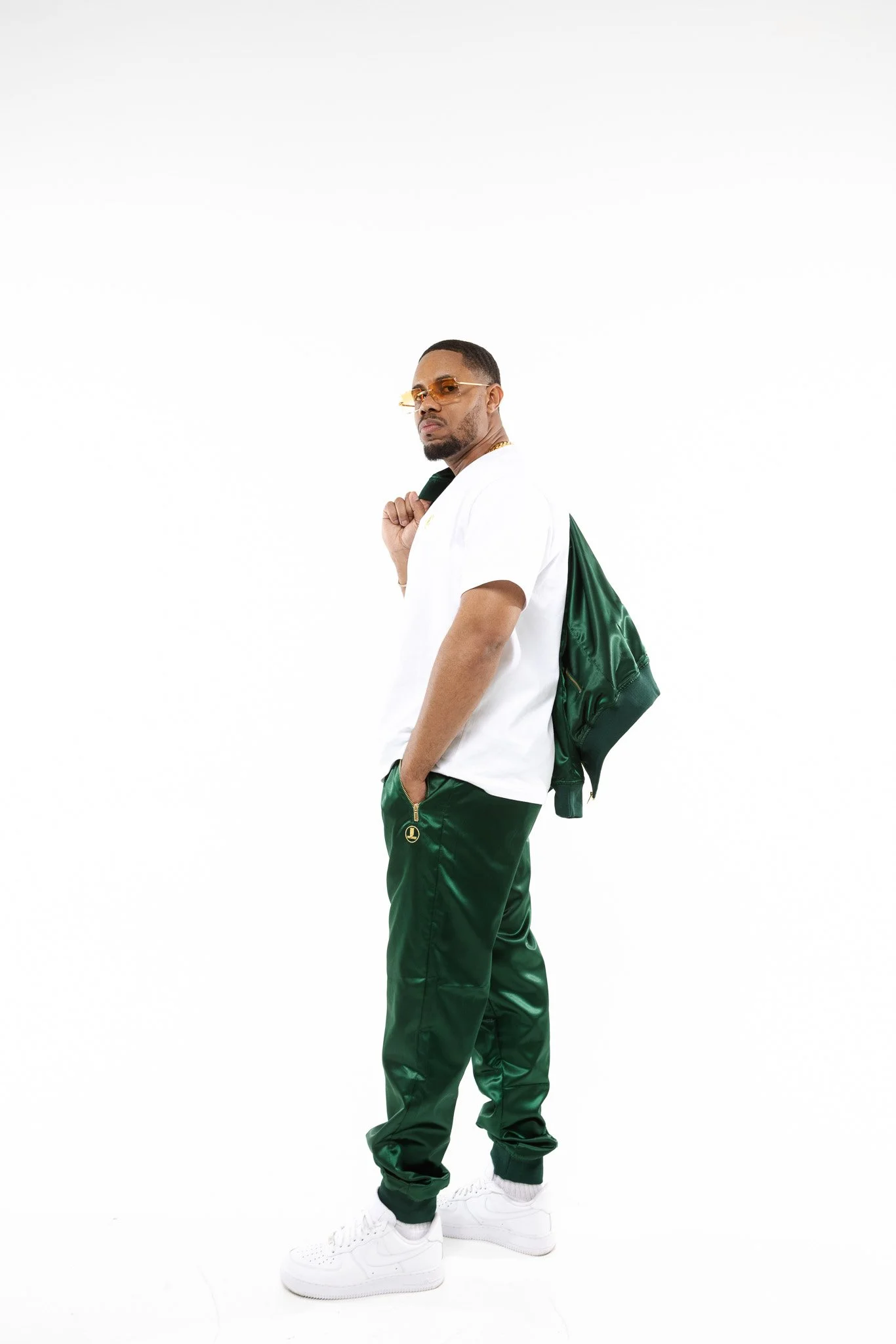 A young man with dark skin wearing orange sunglasses, a white T-shirt, green track pants, and white sneakers, carrying a green jacket over his shoulder, posing against a plain white background.
