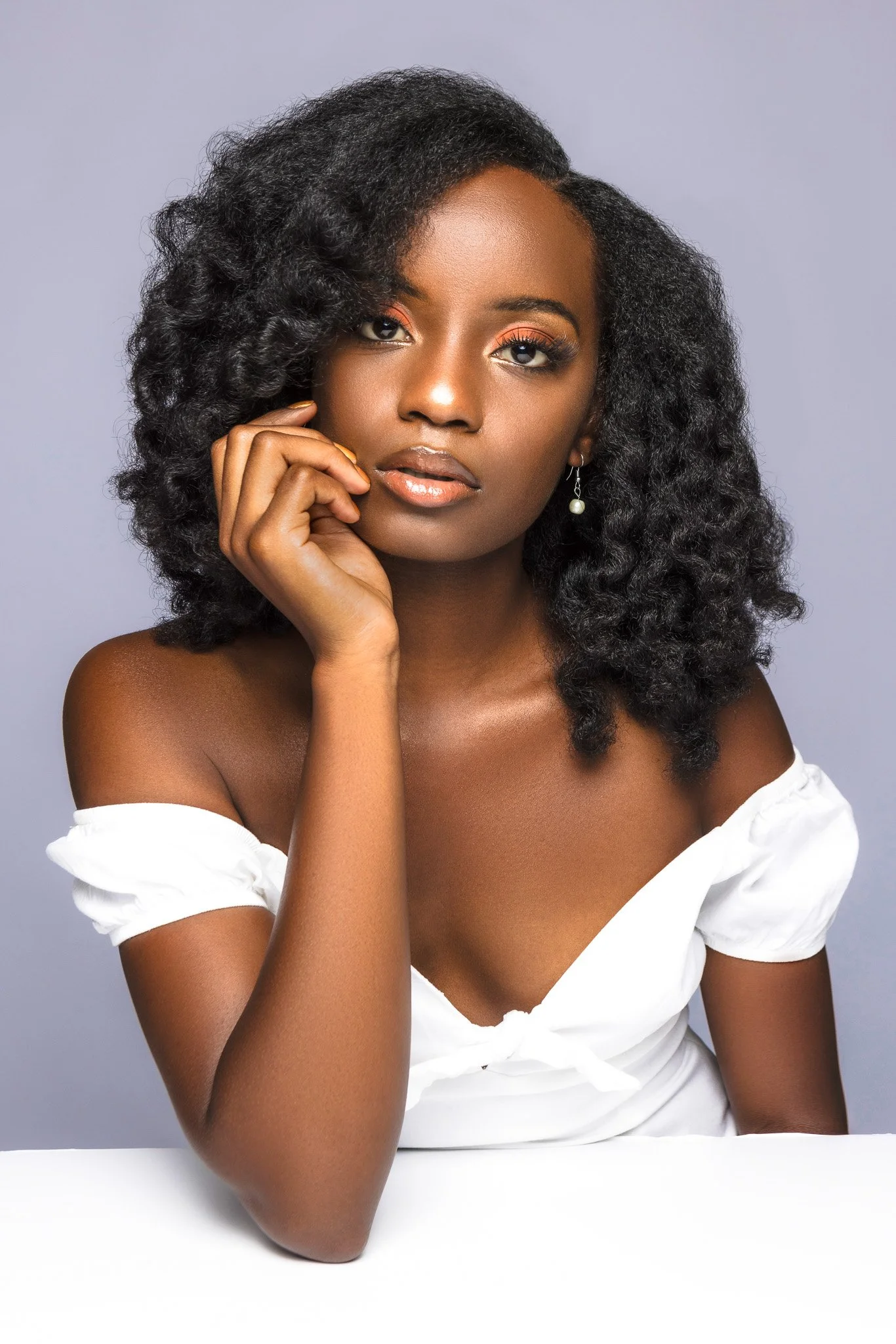 Close-up portrait of a young woman with natural curly hair, wearing a white off-shoulder top, gold hoop earrings, light makeup, and outdoors pose with her hand gently touching her face against a neutral background.