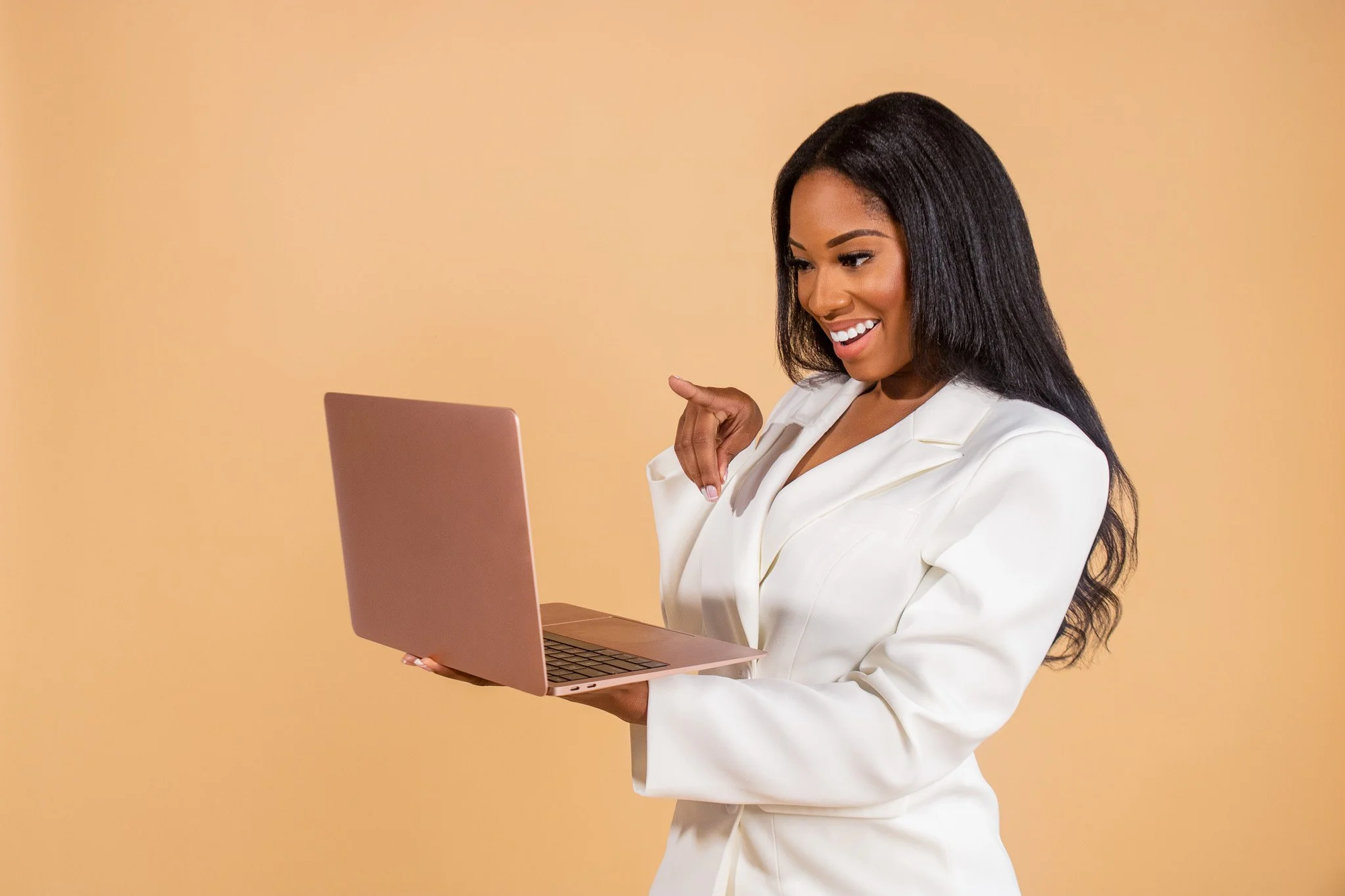 A woman with long black hair wearing a white business suit, holding a pink laptop, smiling and looking at the screen against a plain peach-colored background.