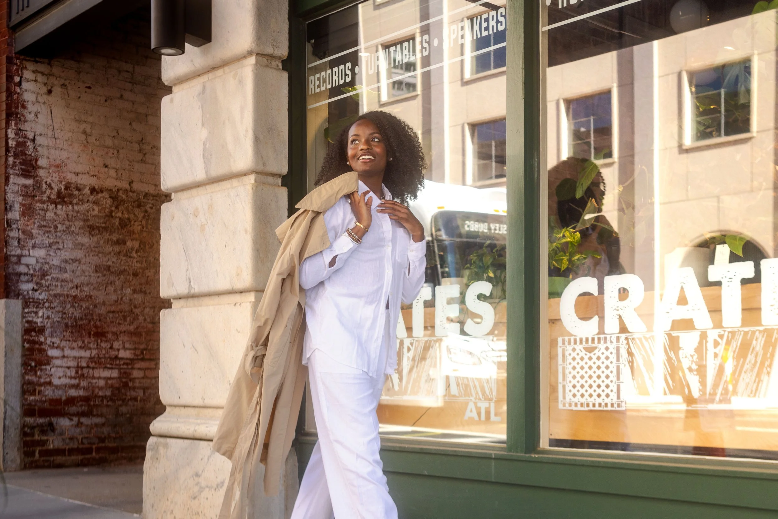 A woman with curly hair wearing a white shirt and white pants standing outside a record store, holding a beige jacket over her shoulder and looking up with a smile. The store window behind her has white text that reads 'RECORDS, TURNTABLES, SPEAKERS' and other partially visible words. The store is located in a brick building with decorative stonework.