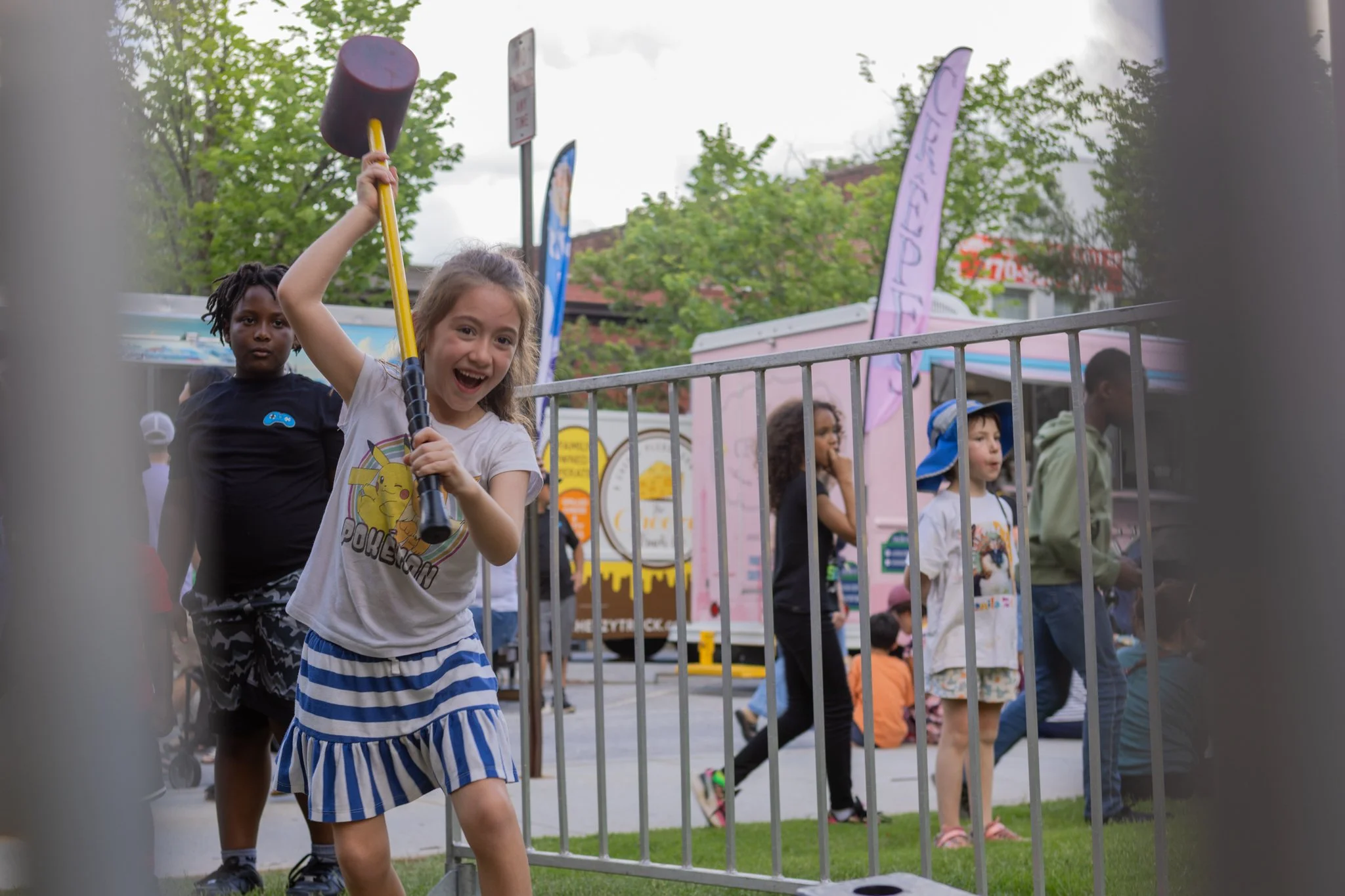 Children attending an outdoor event, with two girls in the foreground. The girl in front has light skin, curly hair, and is smiling while holding a sledgehammer. The girl behind her has darker skin, dreadlocks, and a serious expression. Other childre