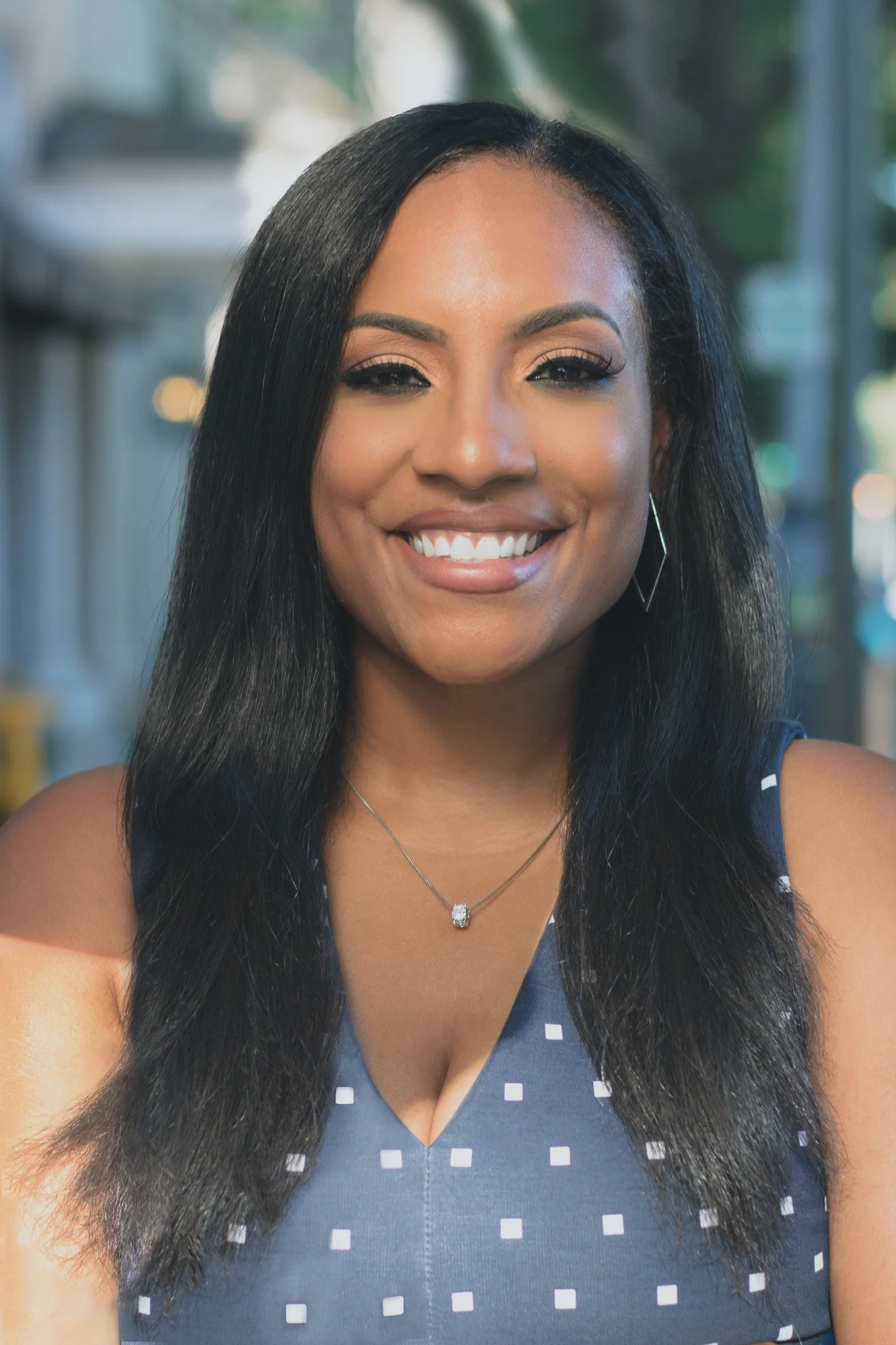 A smiling woman with long black hair, wearing a blue and white checkered dress, jewelry, and makeup, outdoors with a blurred background.