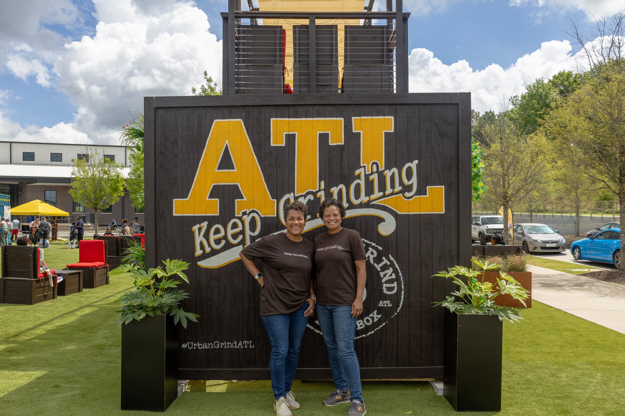 Two women standing in front of a large black sign with yellow and white lettering that reads 'Keep Grinding' and '#UrbanGrindATL'. The women are smiling, dressed in matching black T-shirts and blue jeans, and are outdoors on a sunny day with trees, b