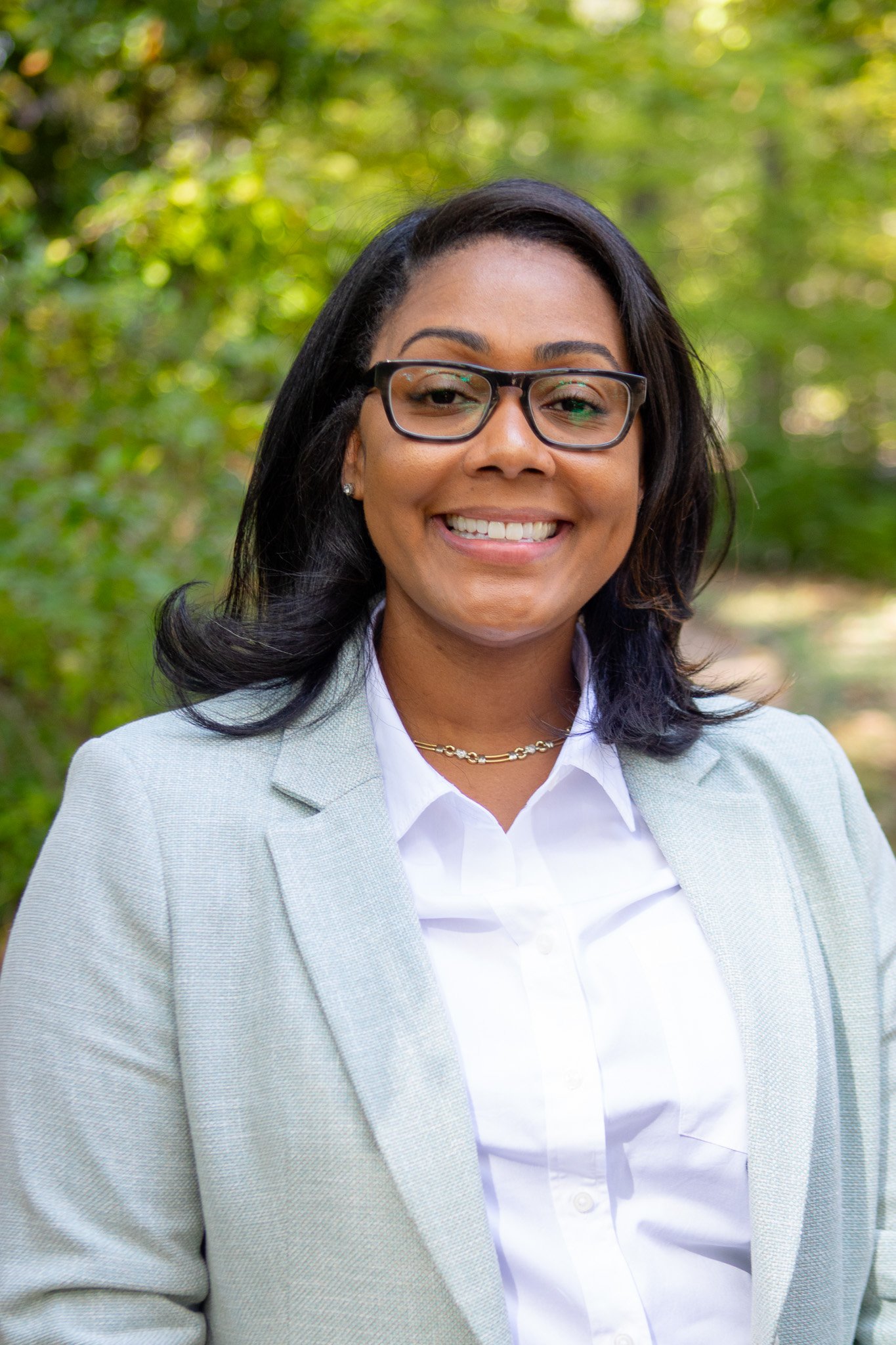 A smiling woman with glasses, wearing a light gray blazer and white shirt, standing outdoors with green trees in the background.