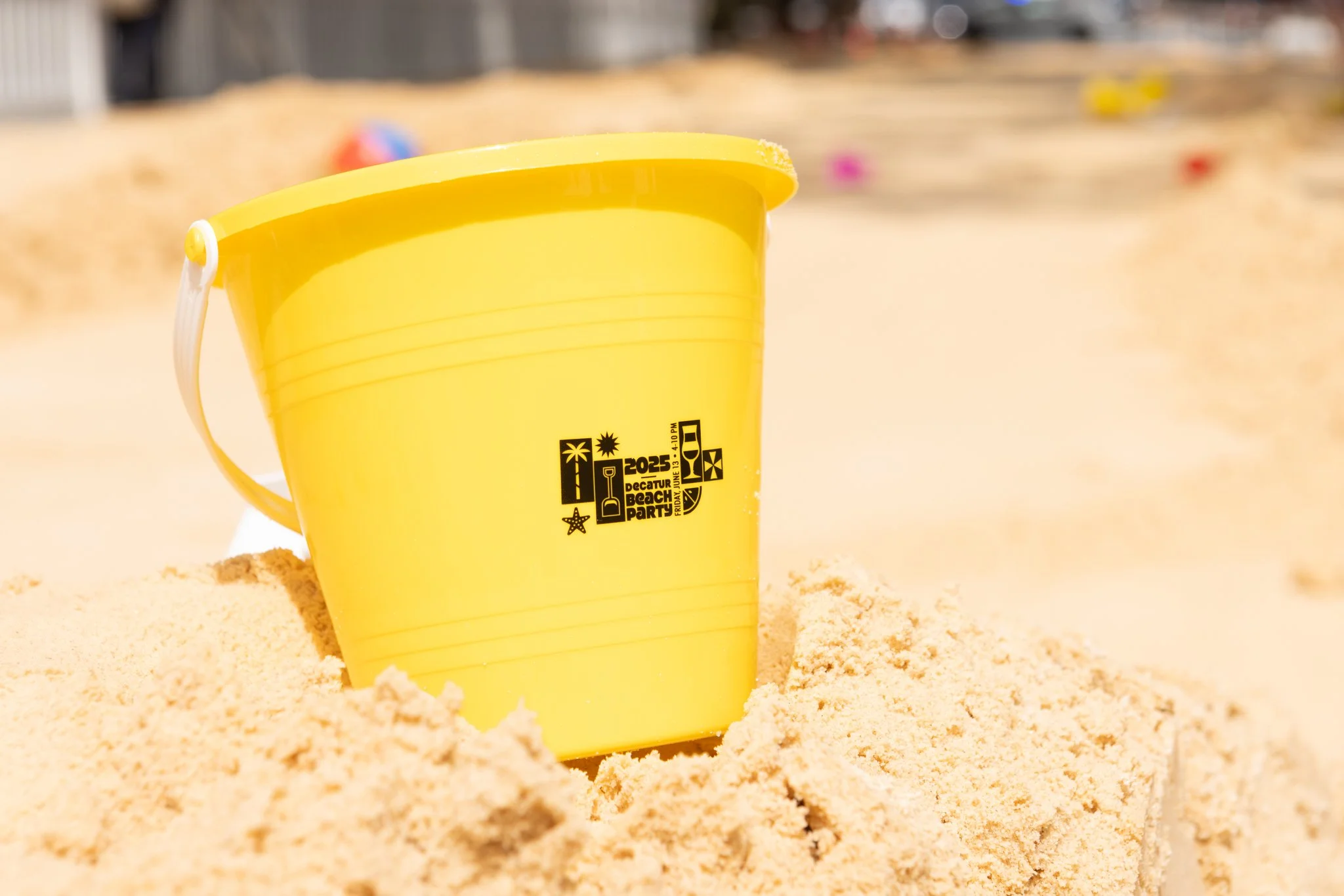 Yellow plastic bucket placed on sandy beach, with colorful objects blurred in the background.