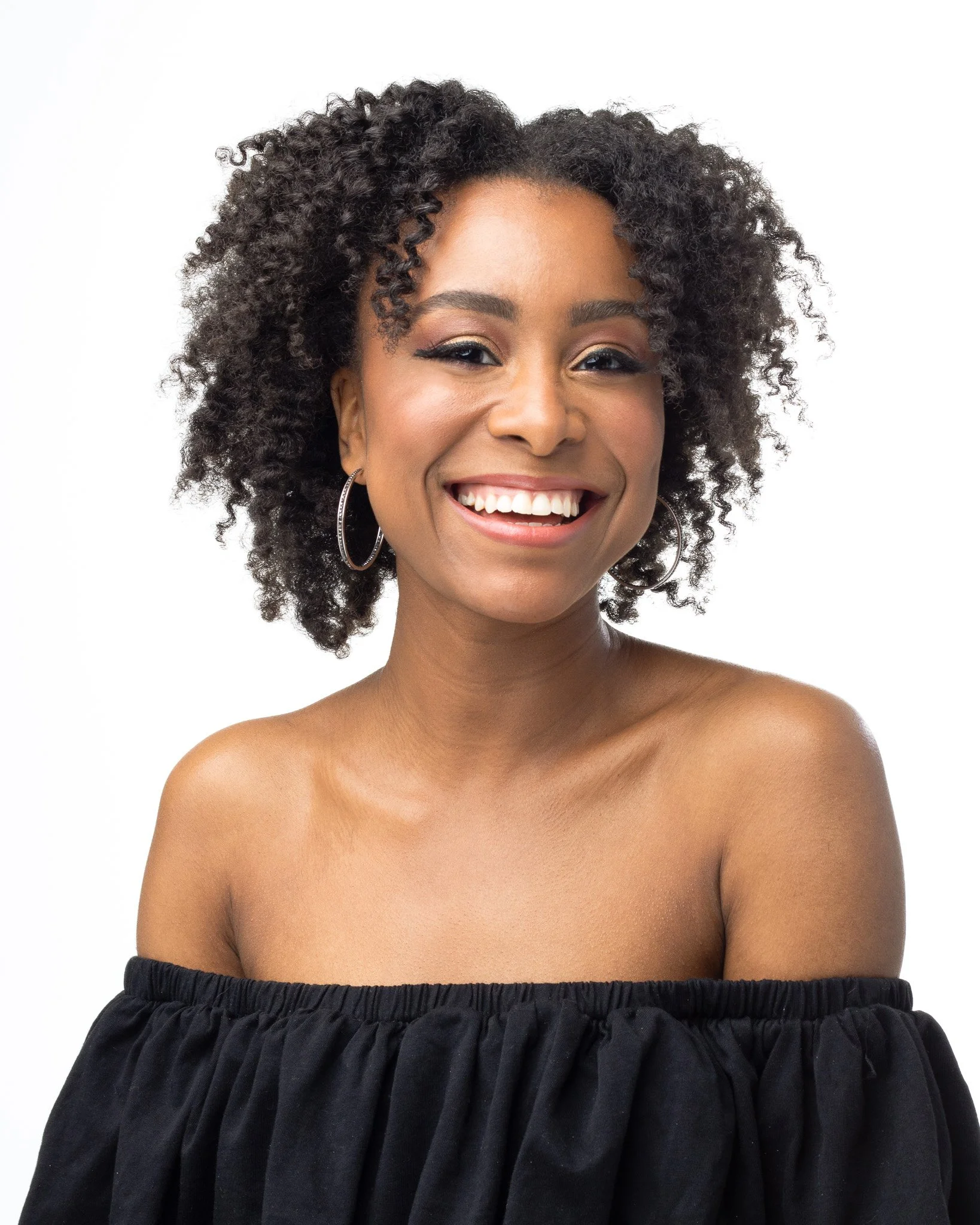 Close-up of a smiling woman with curly black hair, hoop earrings, and makeup, wearing a black off-shoulder top against a plain white background.