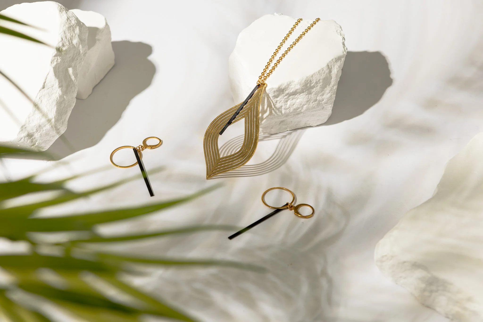 Gold jewelry including a necklace with a leaf-shaped pendant and a pair of earrings resting on white decorative stones with shadows, with green leaves partially visible in the foreground.