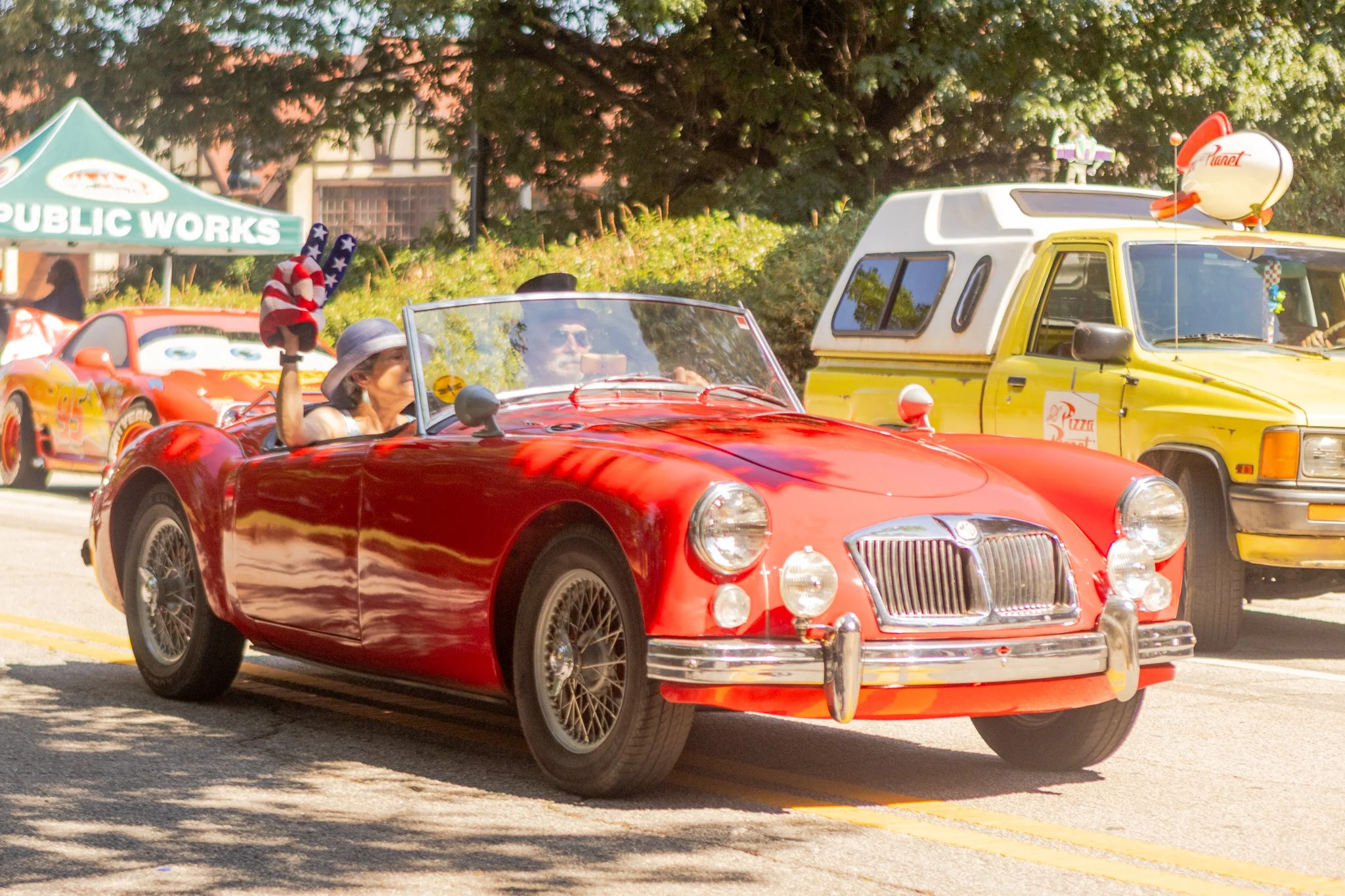A person with a hat and sunglasses driving a red vintage car in a parade or outdoor event, with other vehicles and a green tent in the background.