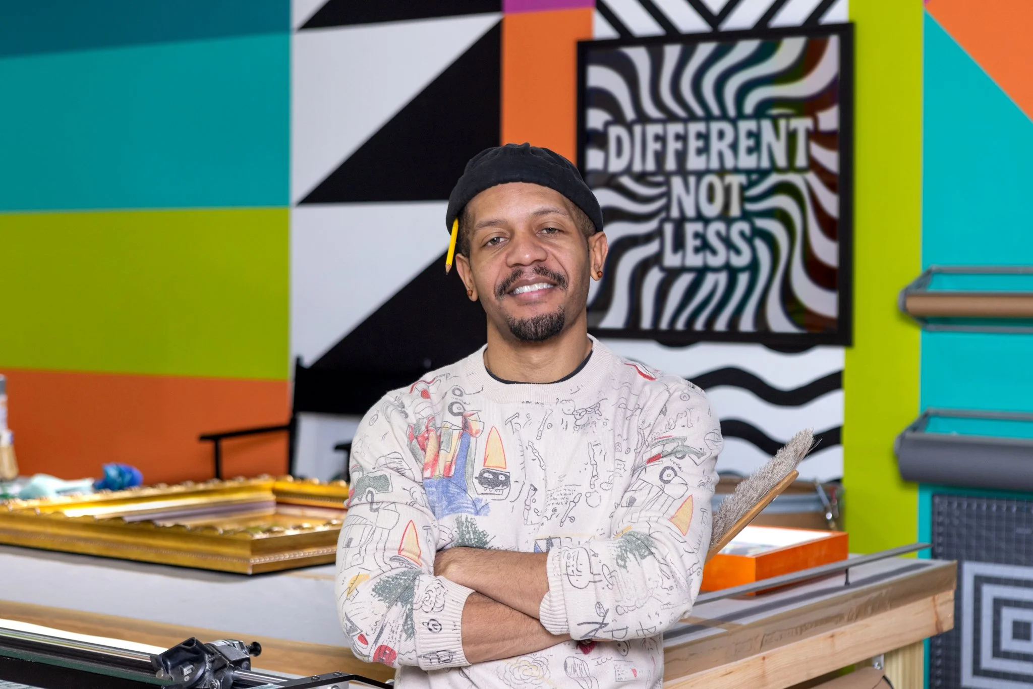 A young man with a black beanie and a themed sweatshirt stands with his arms crossed in a colorful artistic room. The background features bold, geometric and wavy black-and-white patterns, with a sign that says 'Different Not Less' and bright walls i