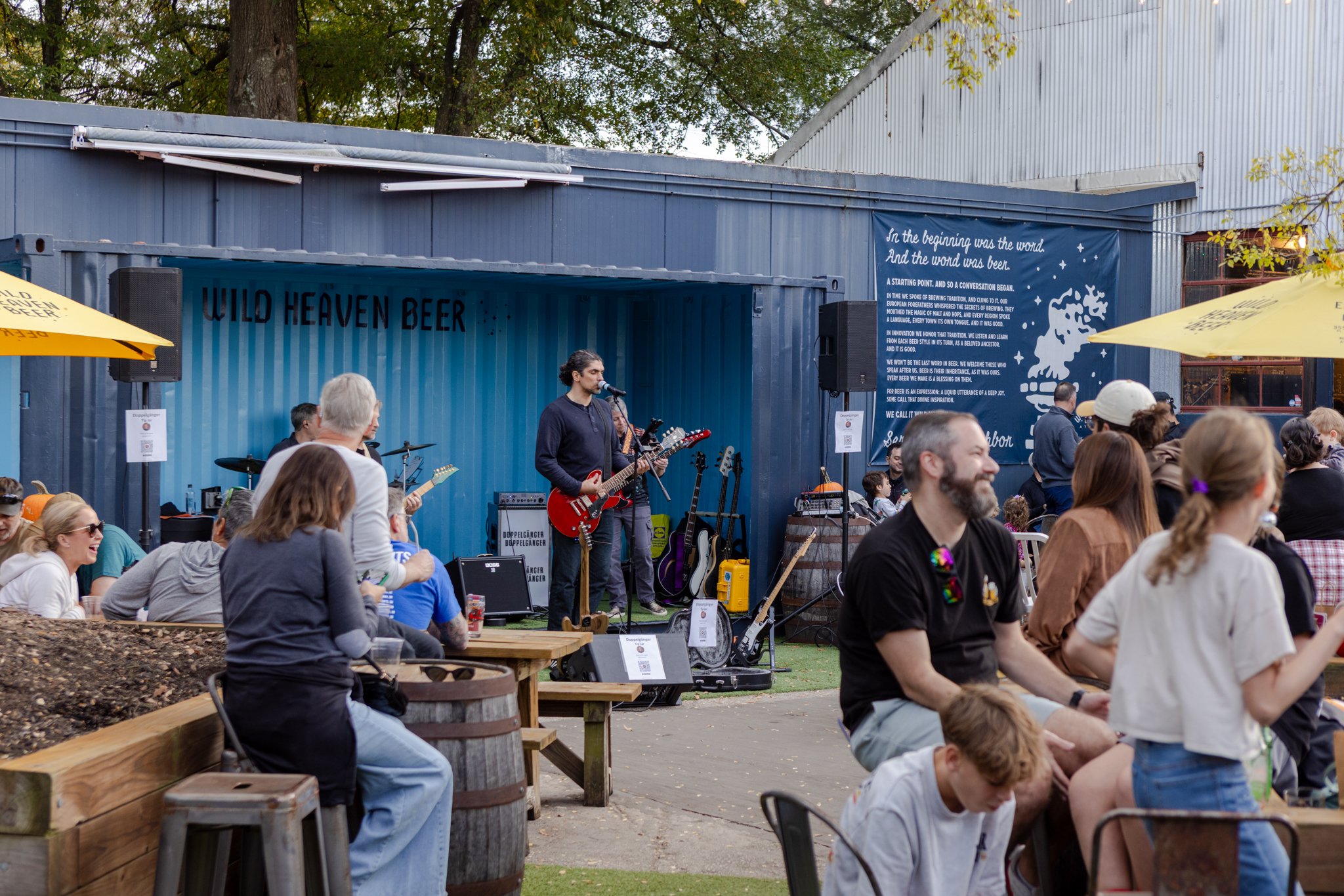 An outdoor gathering where a band is performing on a stage labeled 'Wild Heaven Beer' with a crowd of people watching. The stage is made from a shipping container with a blue banner displaying text about beer and brewing traditions. The audience is s