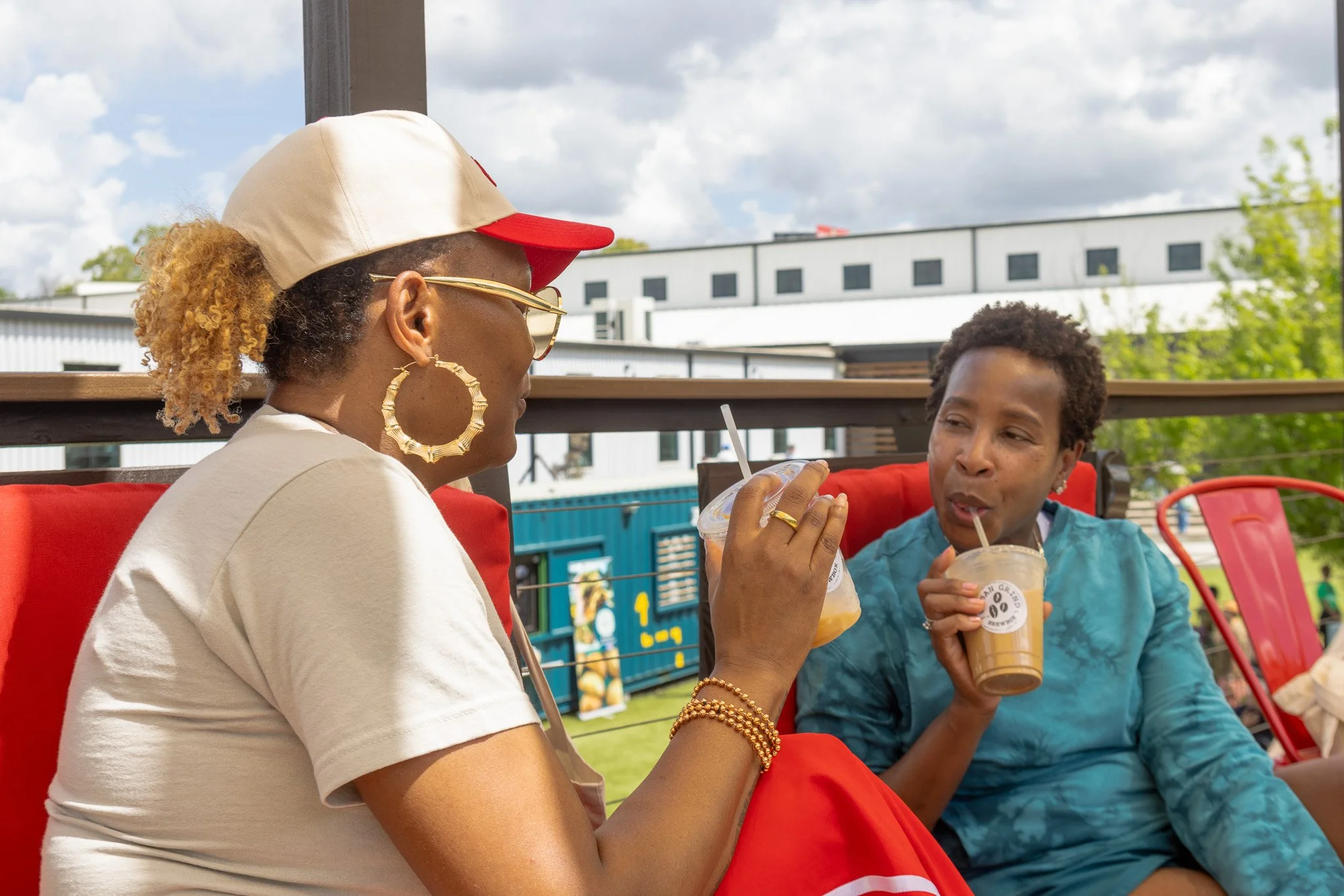 Two women sitting outdoors on red chairs, drinking iced beverages through straws, having a conversation. The woman on the left is wearing a beige and red cap, sunglasses, large hoop earrings, and gold jewelry. The woman on the right is wearing a blue