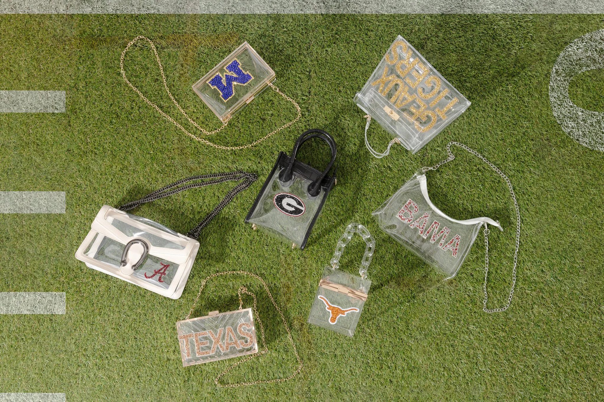 Multiple small, clear handbags with chain straps are arranged on green grass, decorated with university and state names and logos, including Alabama, Texas, Georgia, and others.