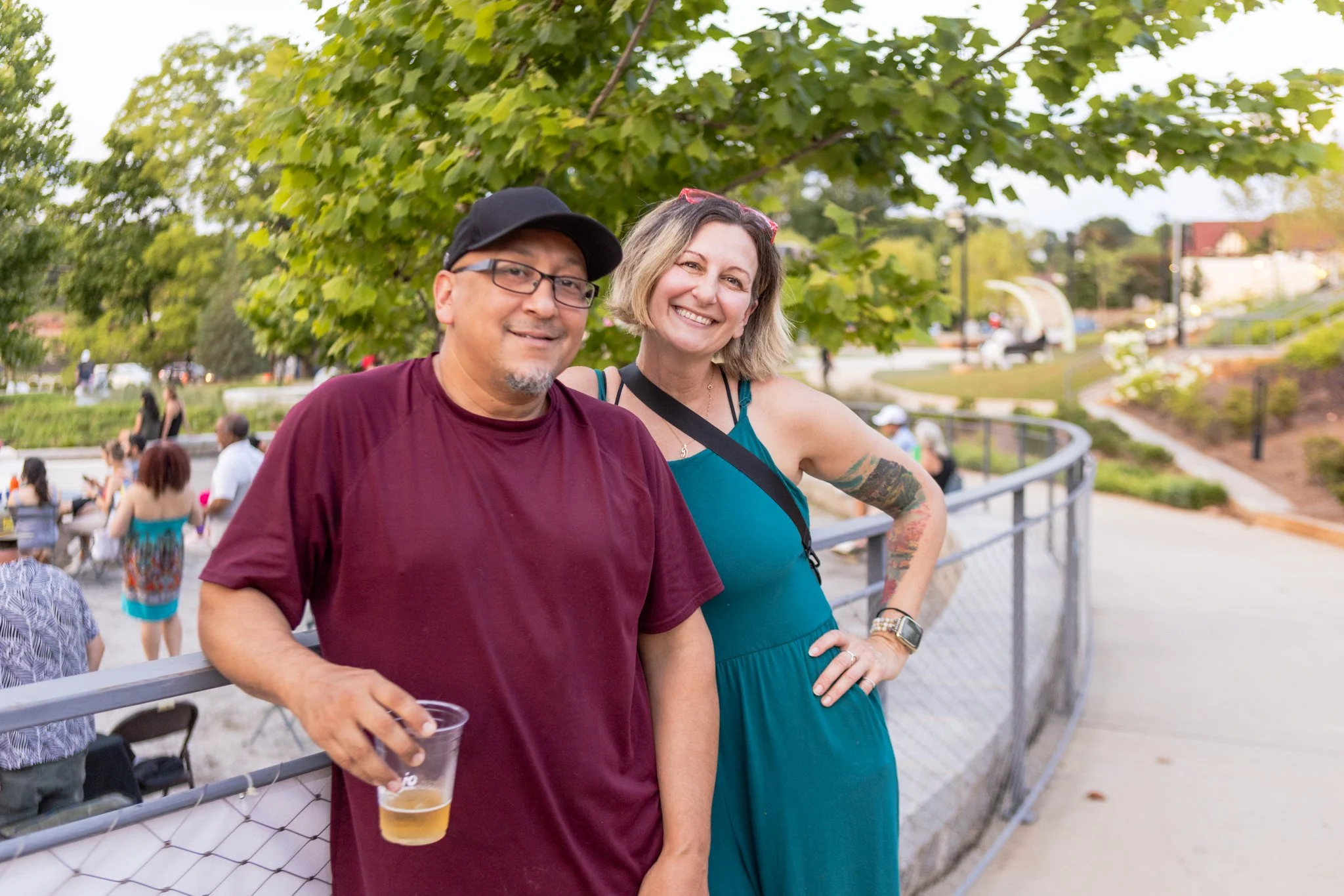 Two smiling adults, a man wearing glasses and a black cap, and a woman with blonde hair and red sunglasses on her head, standing outdoors near a tree with green leaves, with people and a building in the background.
